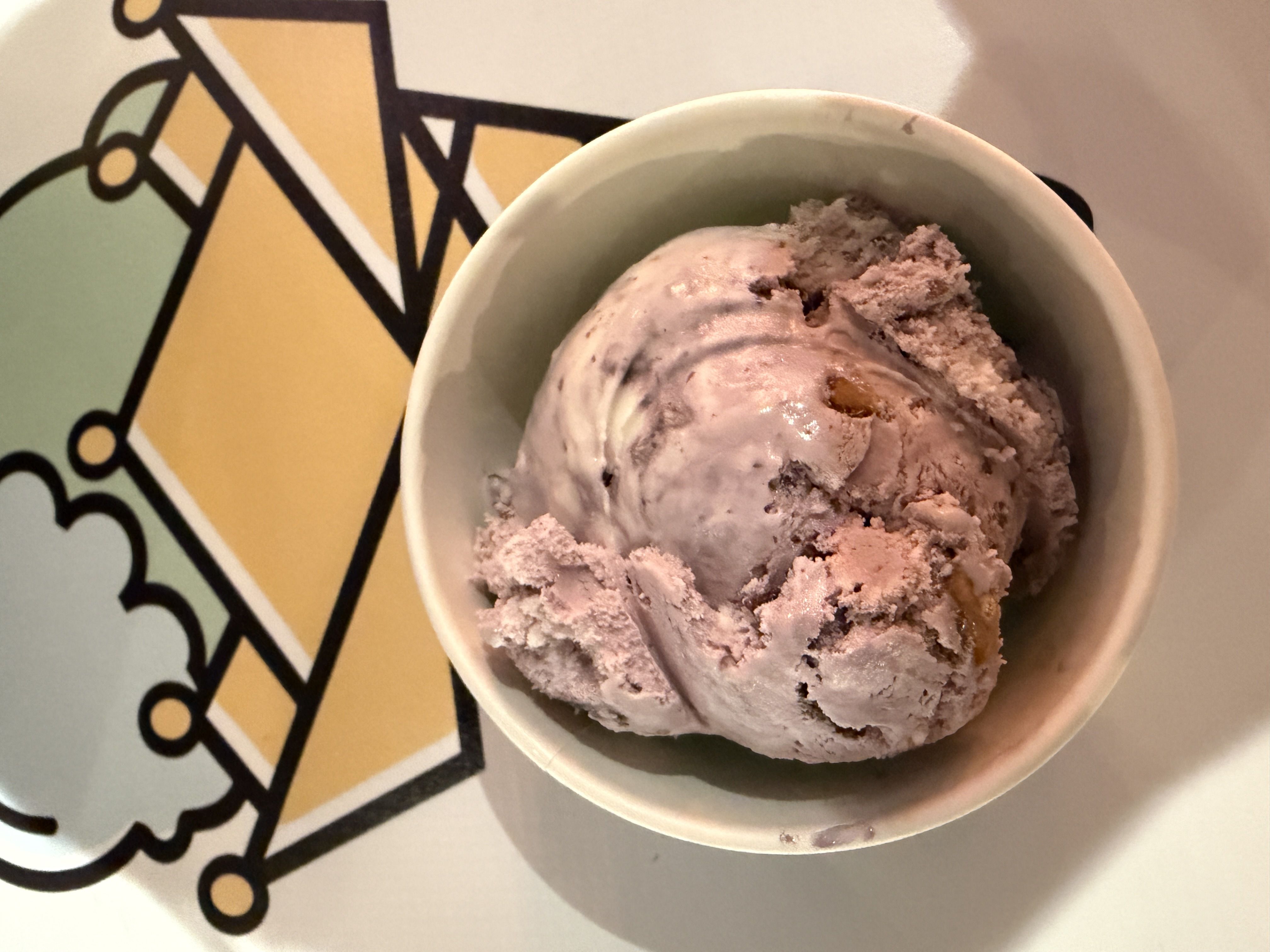 A white bowl of pinkish lavender ice cream with mix-ins, scooped and slightly melted, sits on a colorful geometric background with yellow shapes and bold black outlines.