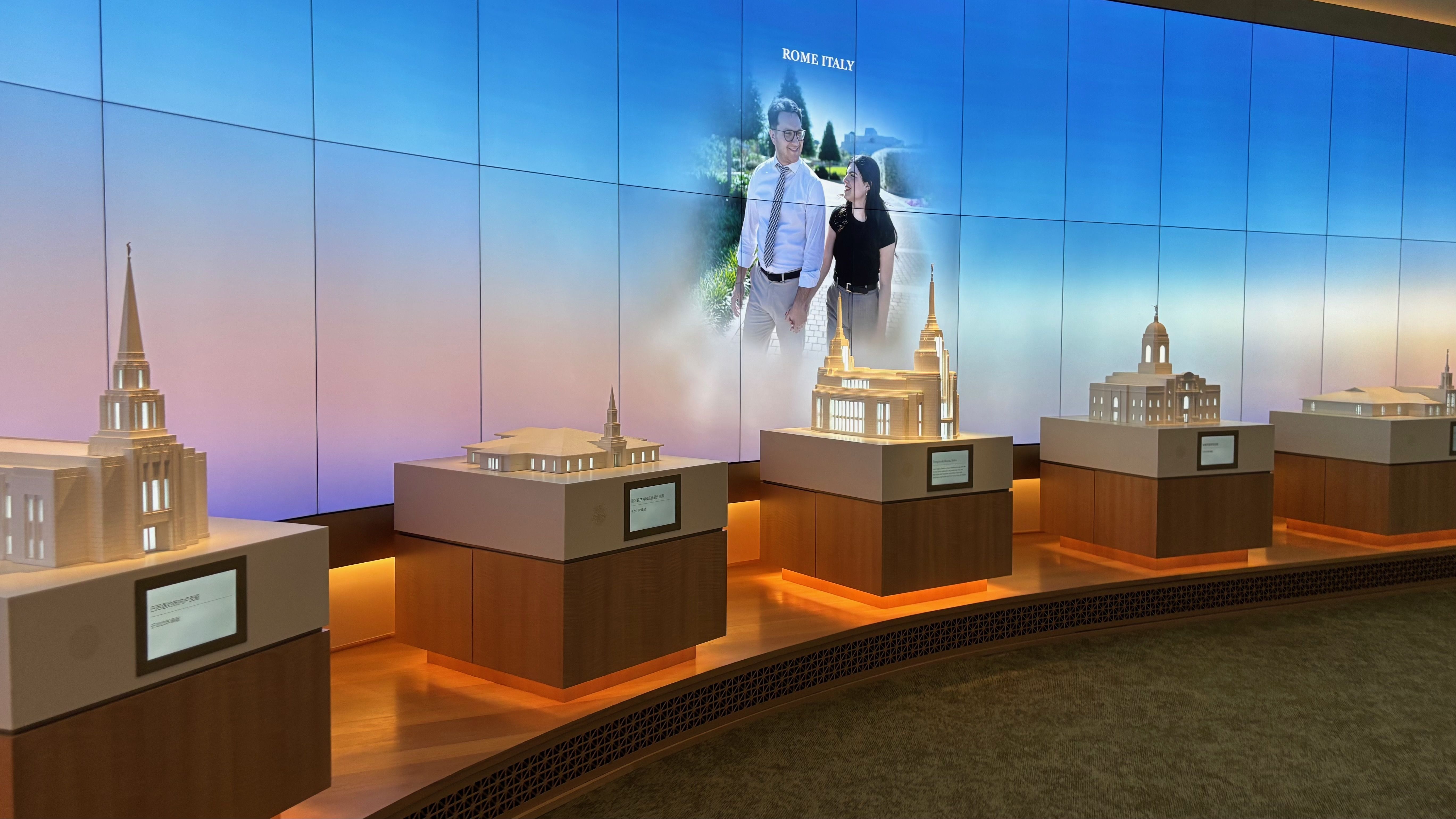 Exhibit of small architectural models on wooden pedestals along a curved platform with orange under-lighting; a blue-to-pink wall mural shows a couple and the words "ROME ITALY" above them.