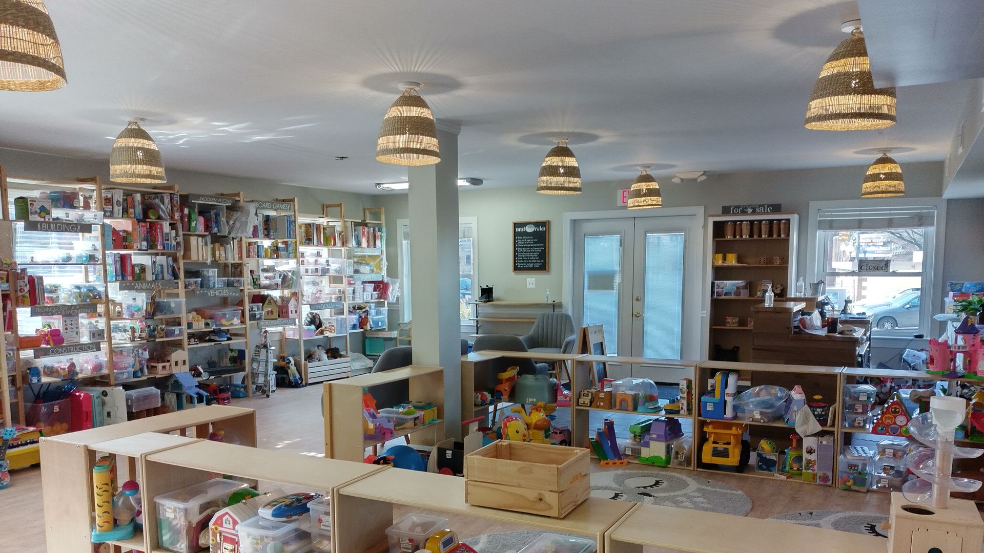 Bright toy store interior with wooden shelves filled with colorful toys, clear plastic bins, and various playthings; woven pendant lamps hang from a light gray ceiling.