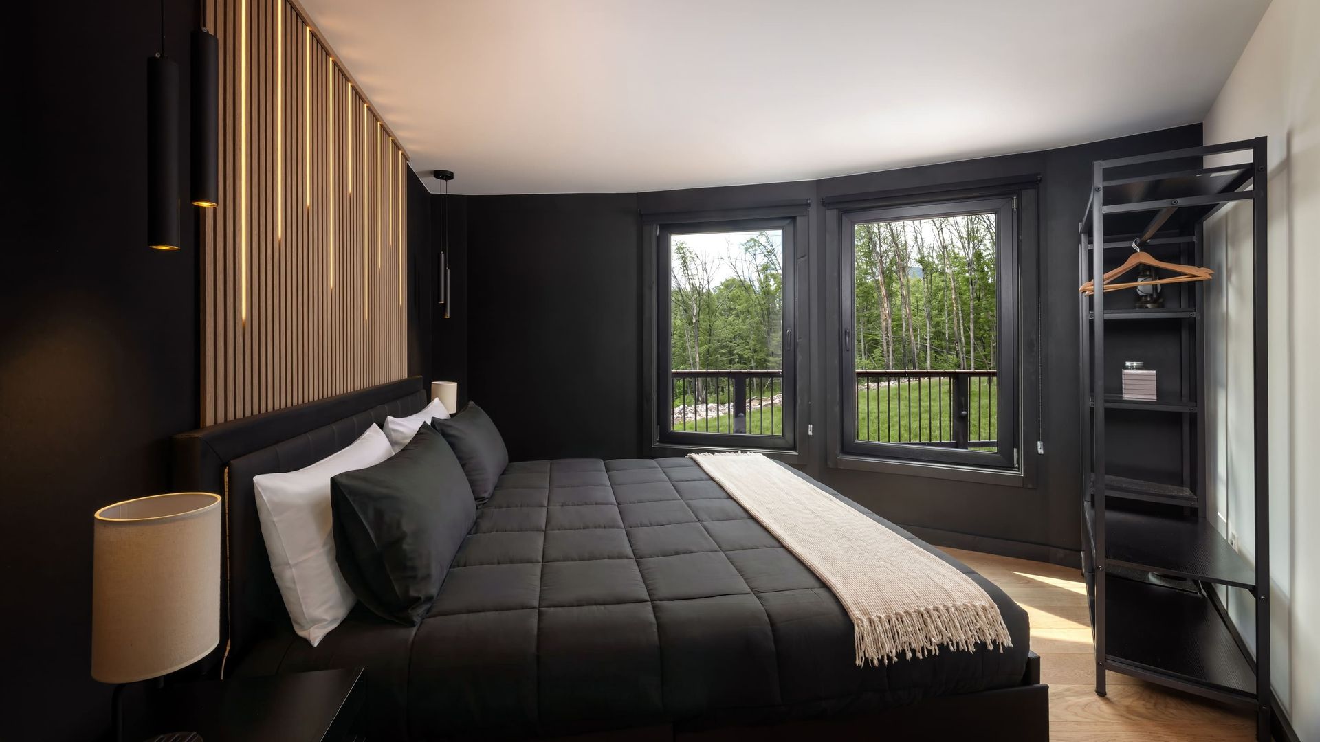 Modern bedroom with black walls and an upholstered black bed with gray bedding. Wooden slat headboard with warm lights. Black-framed windows show trees outside; metal shelf with hangers on the right.
