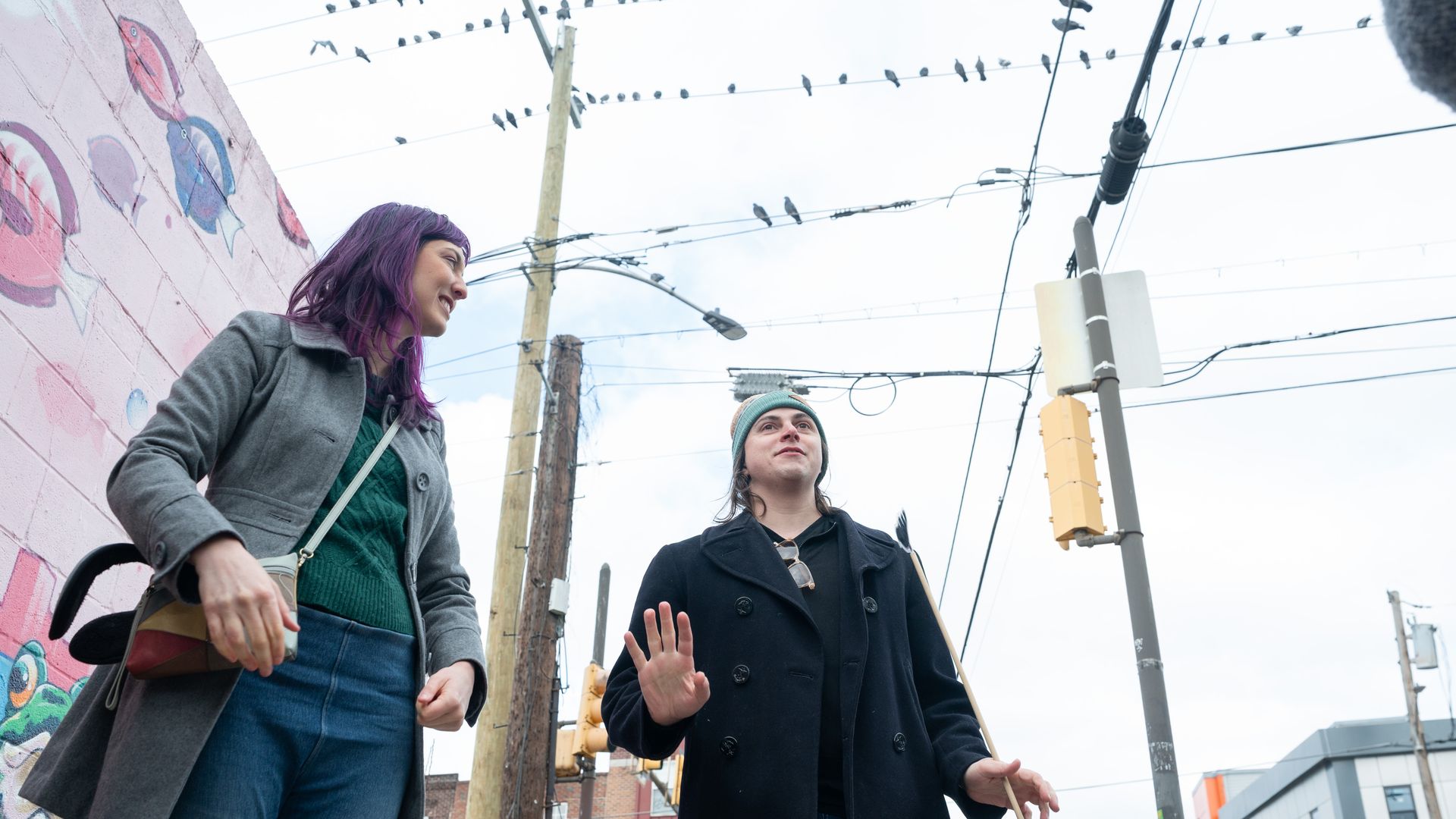 Hannah Michelle Brower, left, and her partner Aspen SImone, walk down a city street beside a pink graffiti wall. Pigeons are perched on overhead power lines.