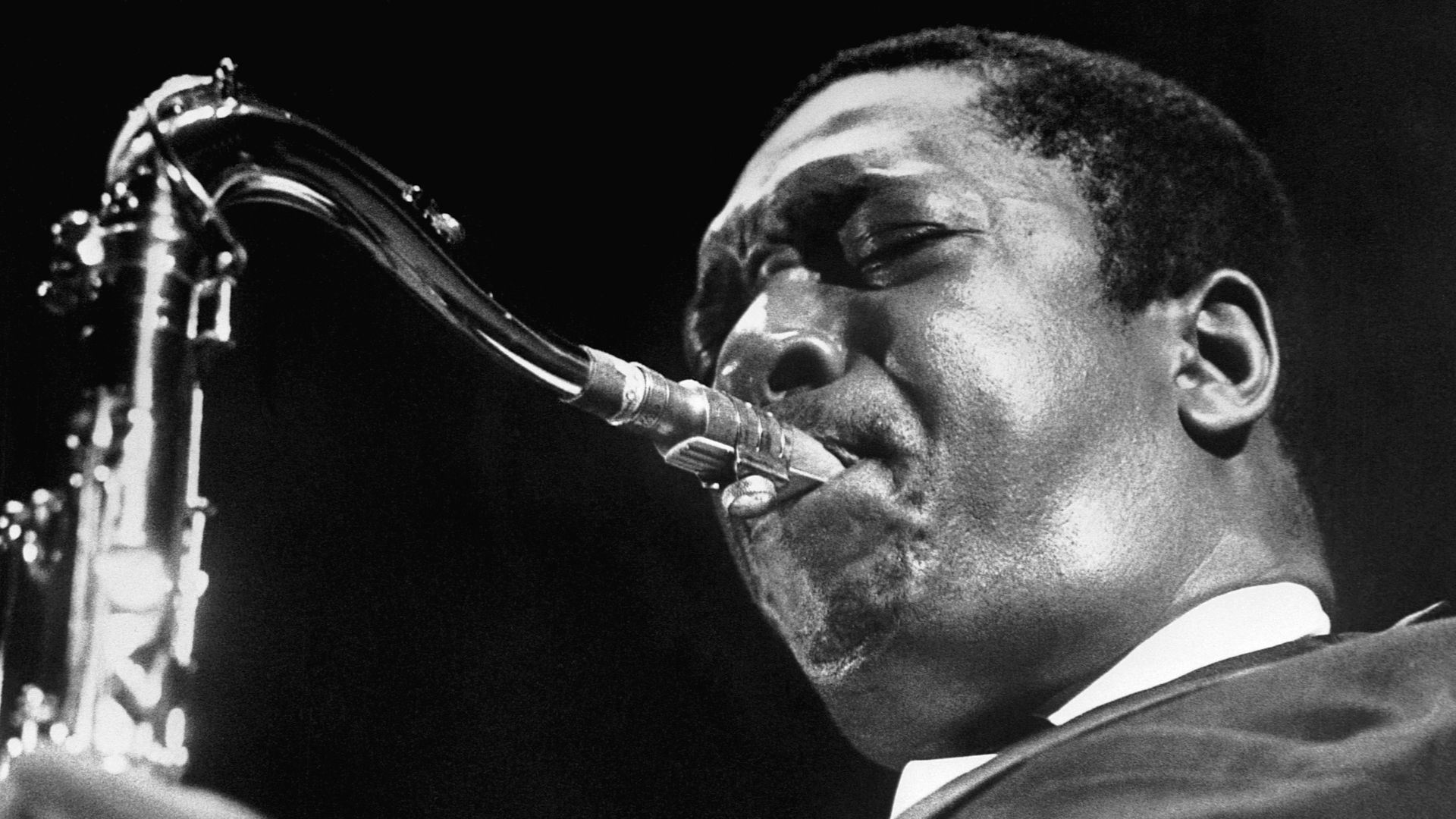 Close-up black-and-white photo of a jazz saxophonist with eyes closed, lips on the mouthpiece, wearing a suit and tie; the saxophone neck and keys appear on the left.