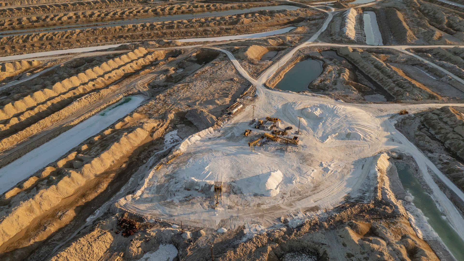 Salt evaporation ponds in California where extraction technology can capture lithium from brine. A large mound of salt-looking material sits in the middle of the frame and is topped with equipment, and trenches with water sit around the mound.