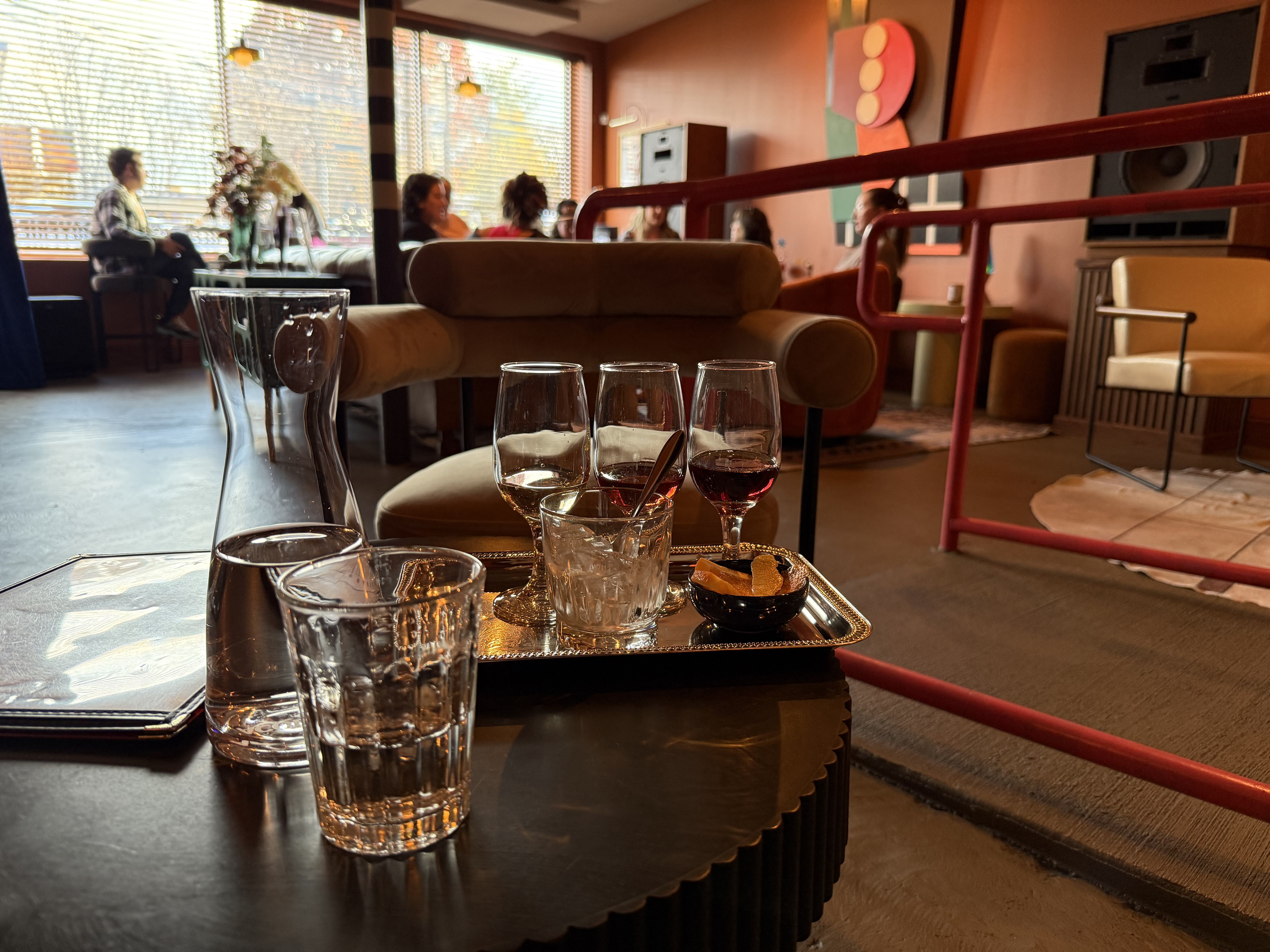Glass carafe and drinking glass on a dark table with a tray holding three wine glasses with different red wines, an ice-filled glass, and a small bowl with orange peels in a cozy lounge.