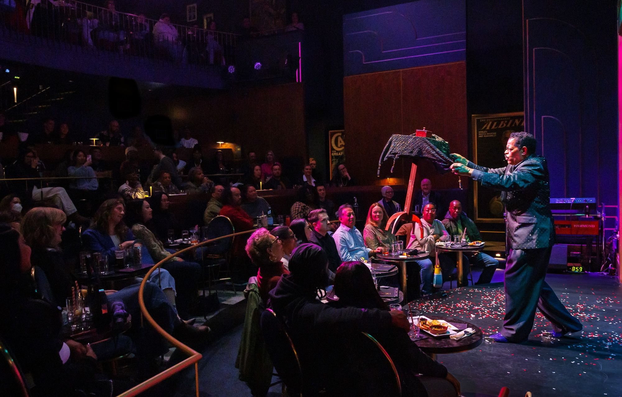A male entertainer in a glittery blue suit performs on a lit stage in a dark club, reaching toward a covered prop as a seated audience watches; purple lighting and confetti dot the floor.