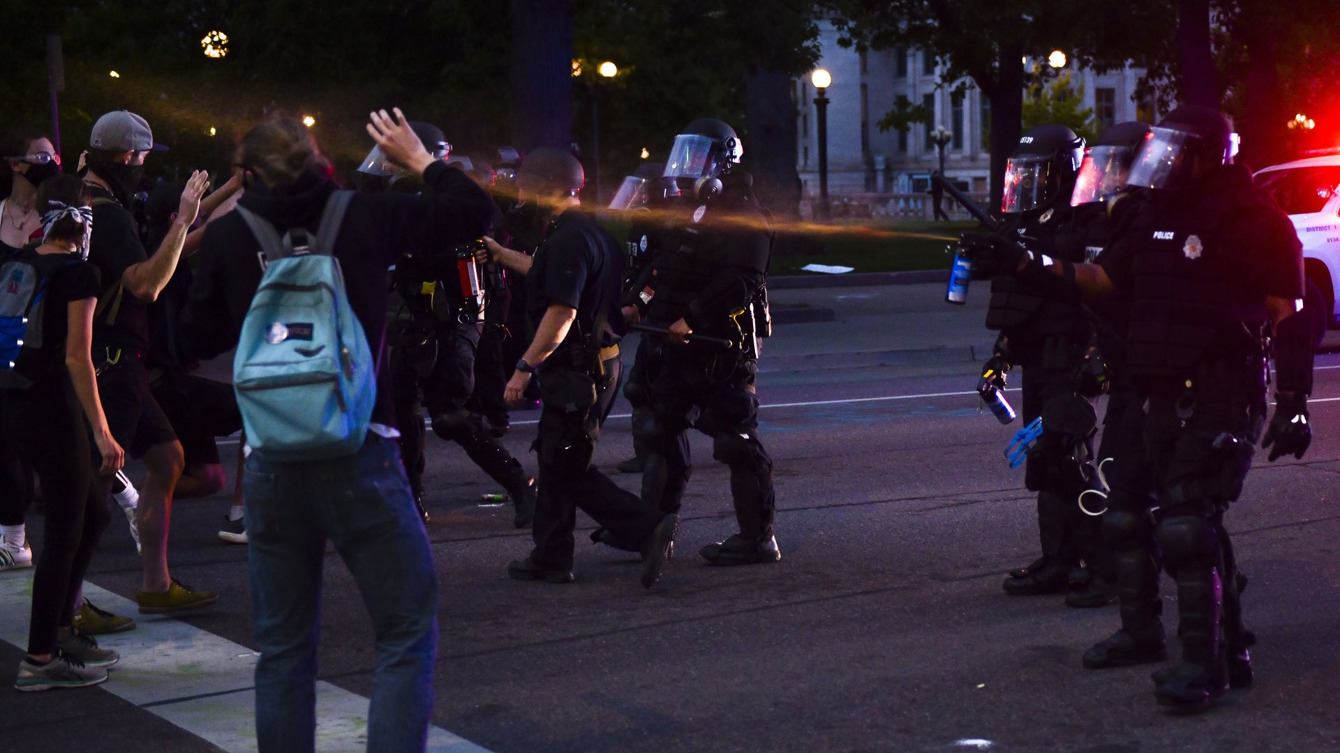 Police in riot gear including plastic masks spray a man with pepper spray. 