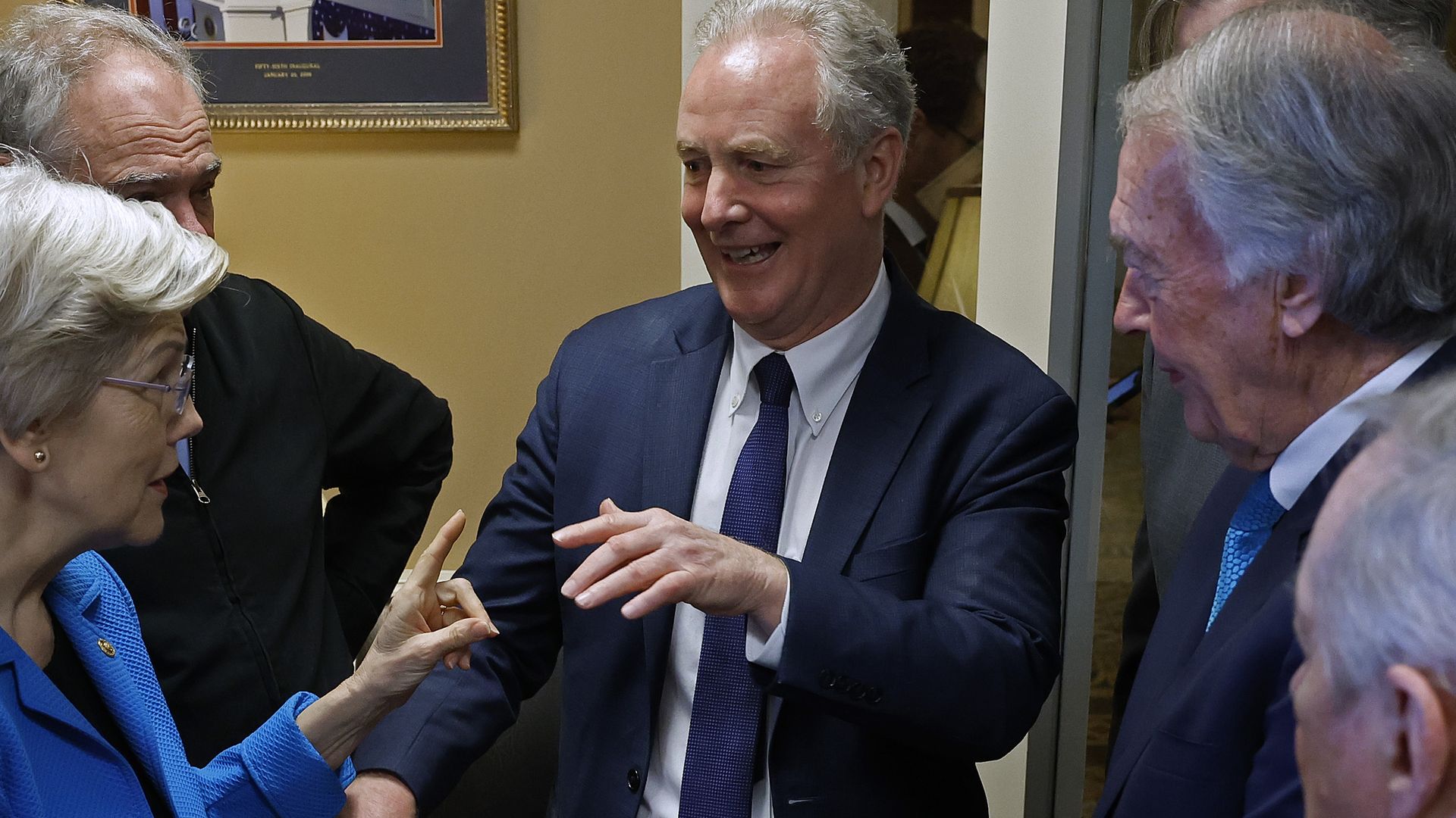 Various people in business suits, mostly men, are chatting in a circle, with one man in the center of the conversation.