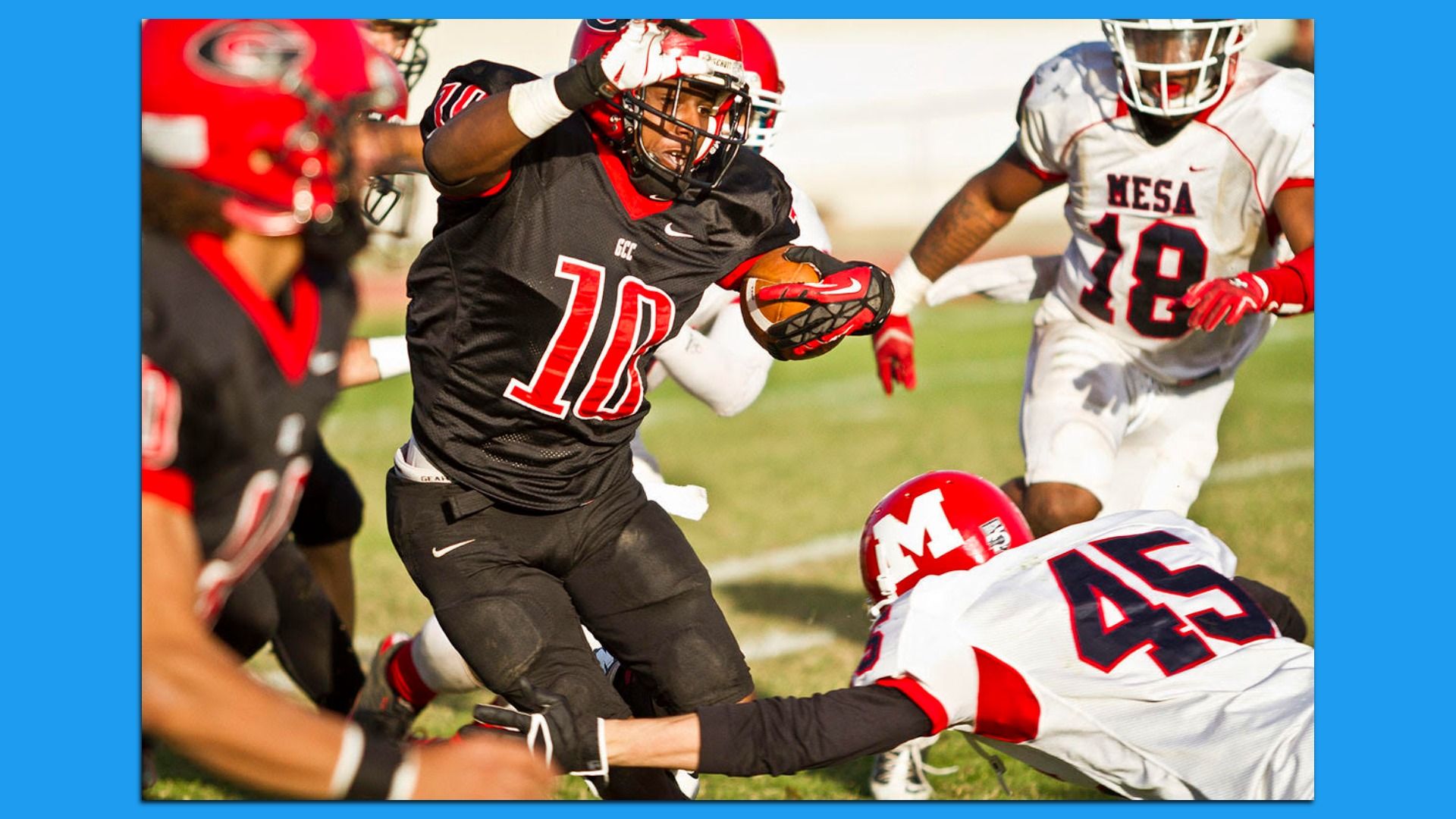 A football player in a red helmet and black uniform with a red number 10 on the chest carries the ball while chased by opponents in red helmets and white jerseys with dark blue numbers on a grassy field.