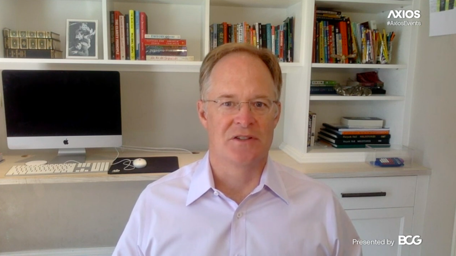 A screenshot of a man wearing a collared shirt and glasses sitting in front of a bookshelf 
