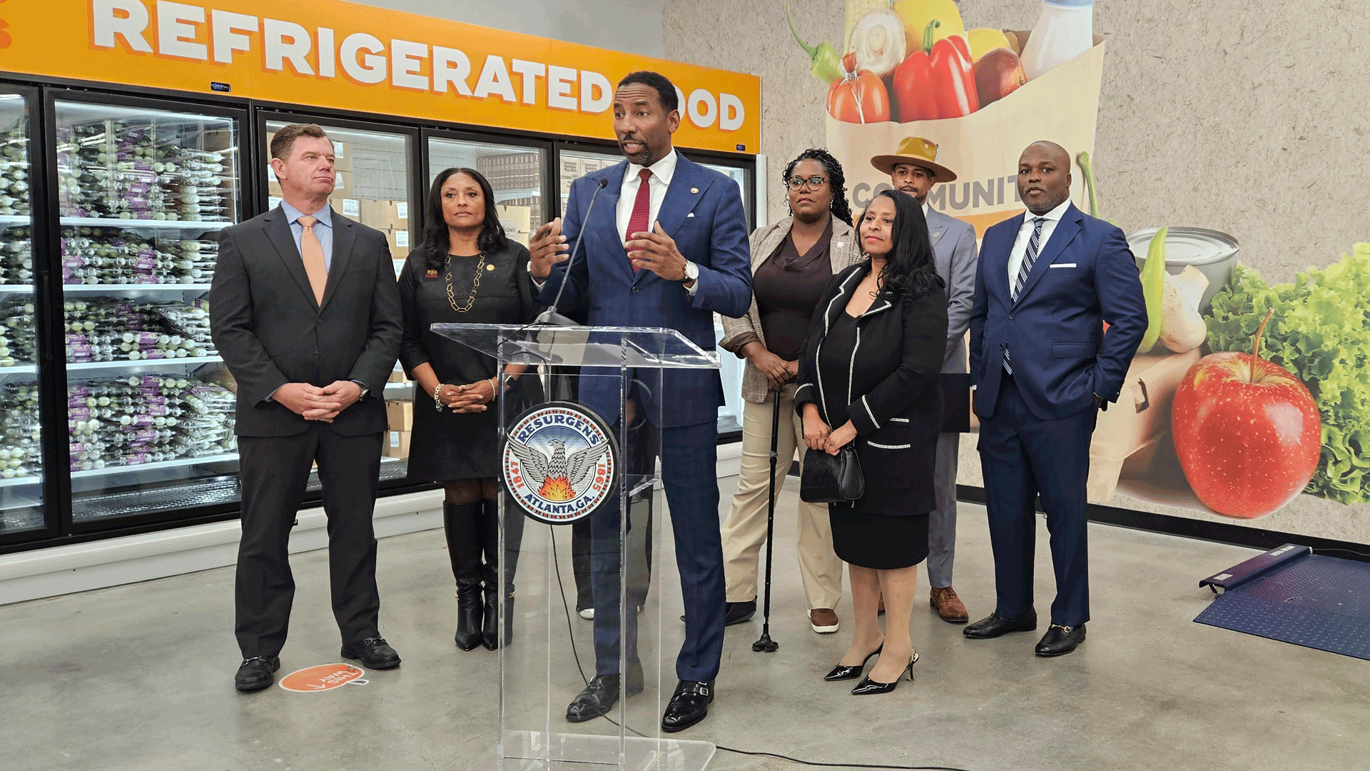 Seven people in business attire stand in front of a refrigerated food section while a man in a blue suit speaks at a clear podium with the city of Atlanta seal. Large produce images are seen on the wall in the background 