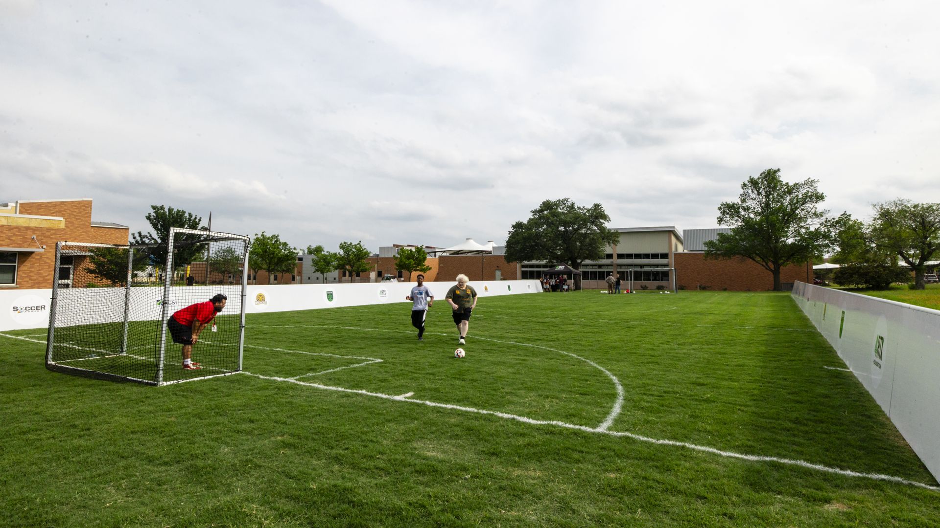 Wide view of a grassy soccer field with a small netted goal on the left, a red-jersey goalie near it, two runners, a white sponsor barrier, brick buildings, trees, and a cloudy blue sky.