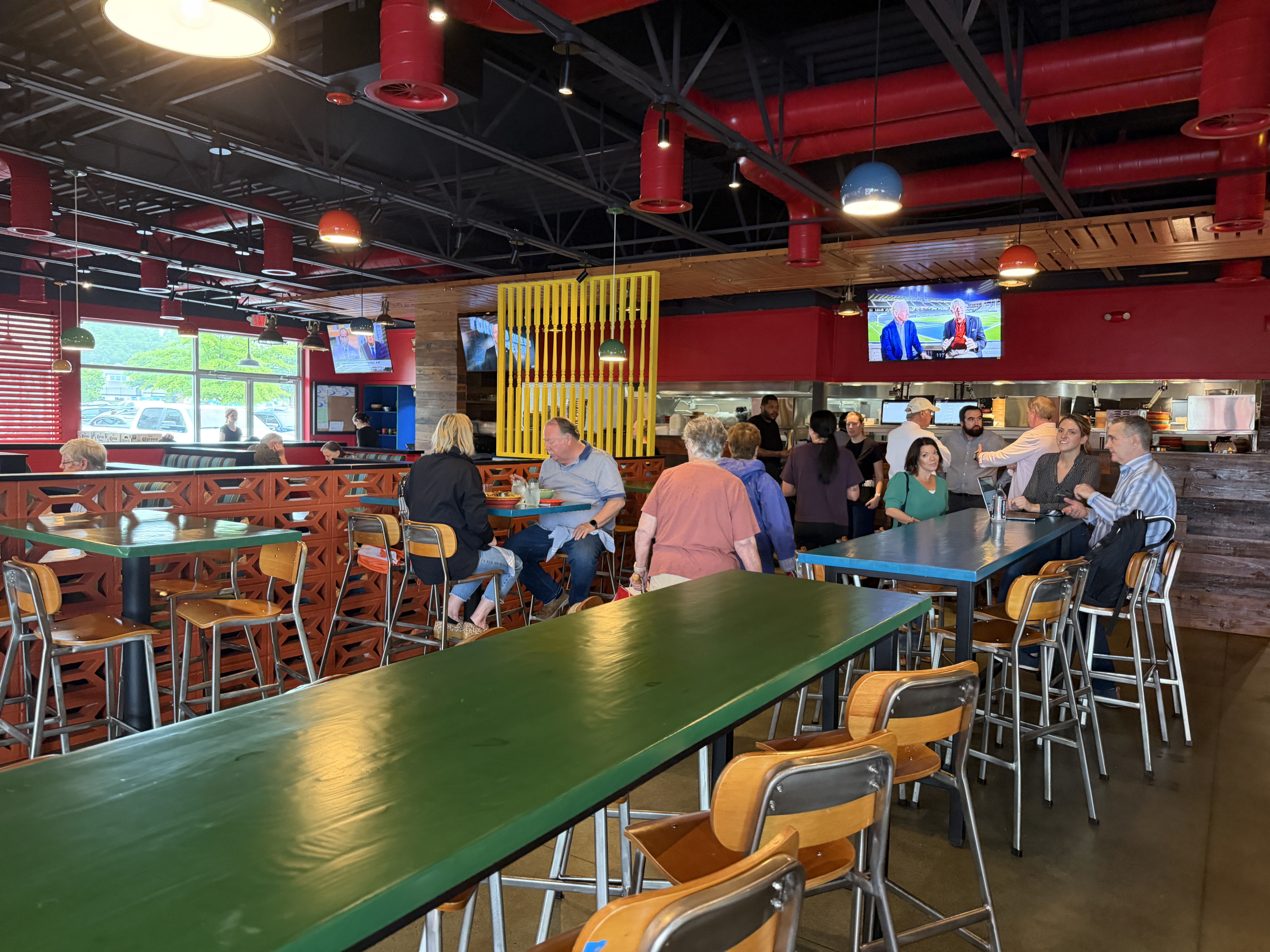 Inside a busy casual restaurant with red walls and exposed ceiling pipes, dark beams, and hanging lamps. Green tables, wooden chairs, a yellow lattice divider, and many diners; TVs show a sports broadcast.