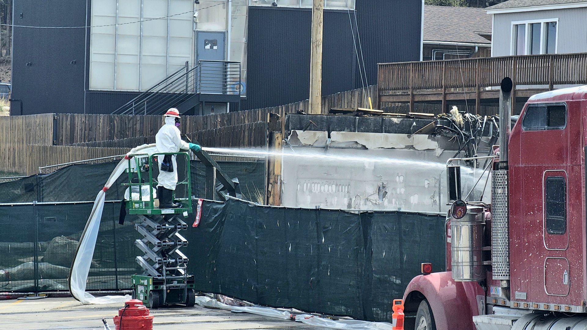 Worker in a white hazmat suit and mask stands on a green scissor lift, spraying water at a damaged concrete wall behind a black construction fence; a red semi-truck is parked to the right.