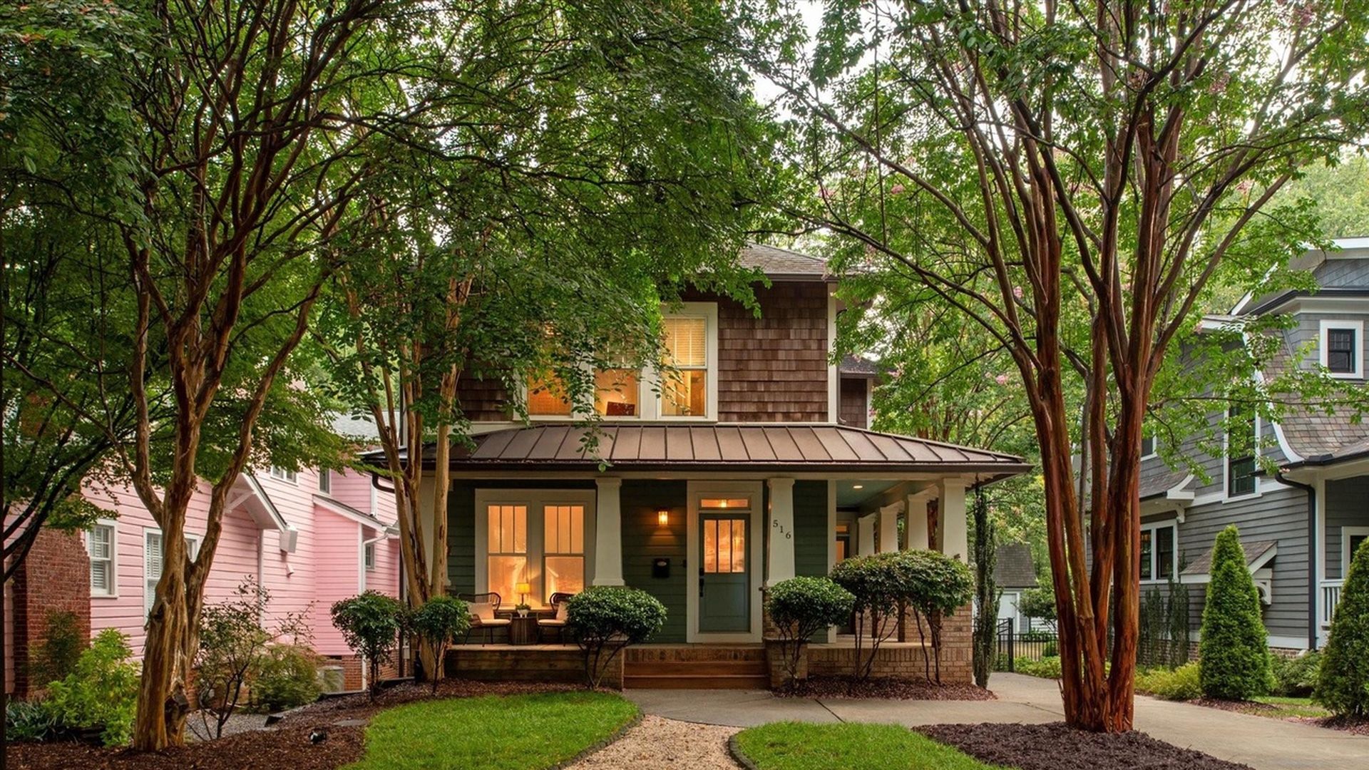 Cozy two-story house with illuminated windows and green front door, surrounded by tall trees, shrubs, and a green lawn with a gravel path leading to the porch.