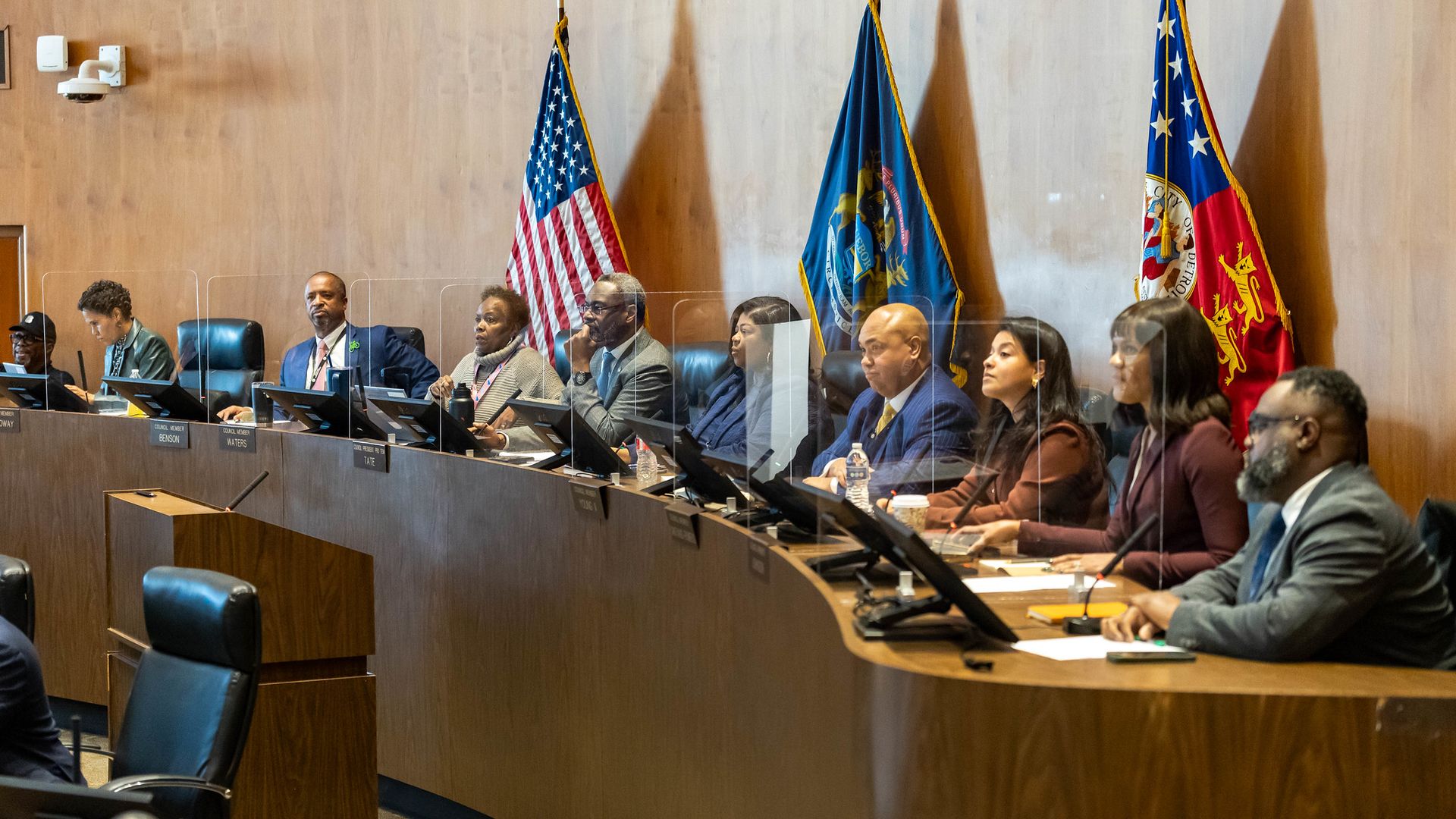 Detroit city council members are seated in their chamber, each at a computer, with flags in the background.