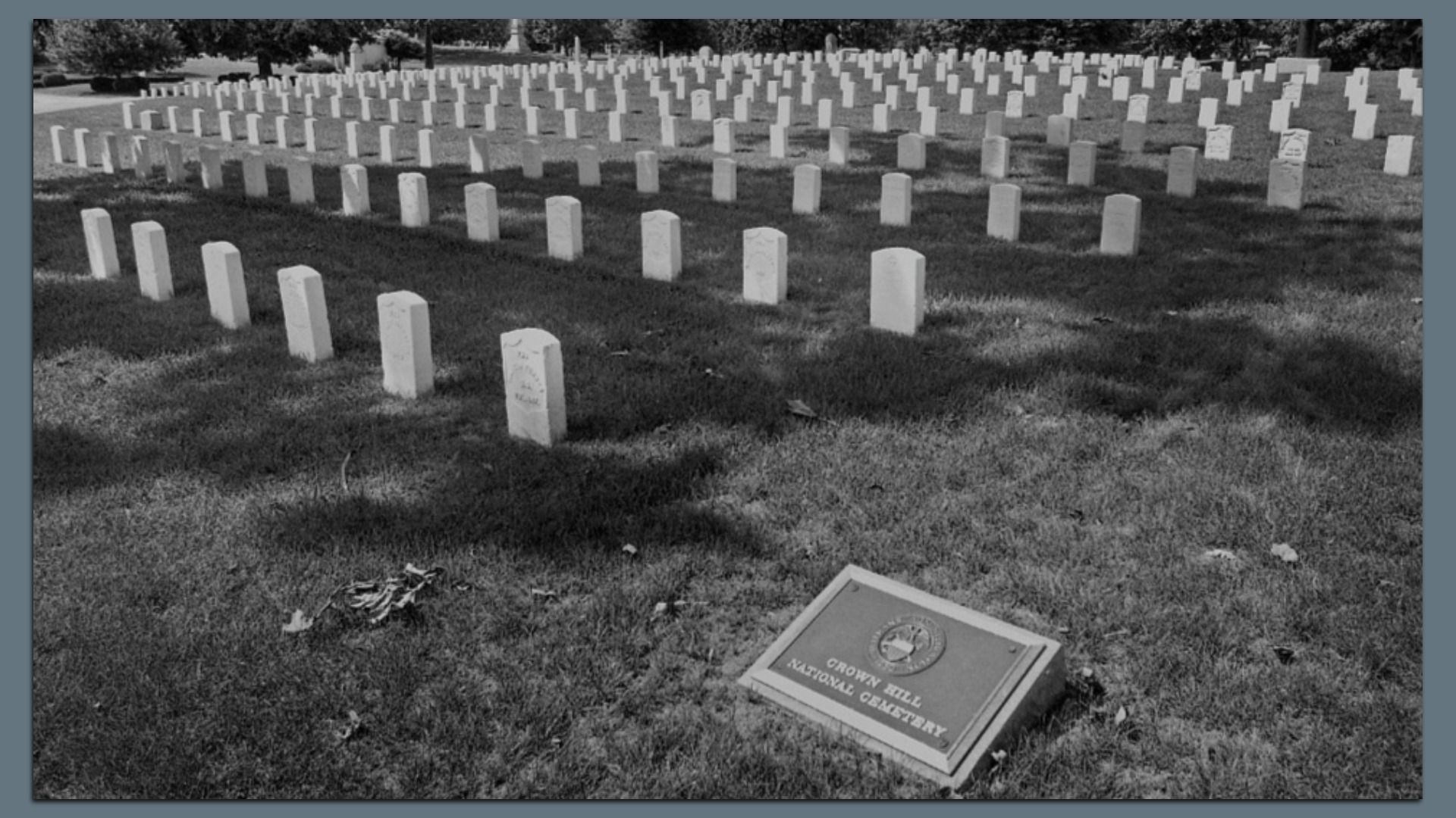 Black and white image of evenly spaced white gravestones in rows on grass, with a plaque that reads "Crown Hill National Cemetery" in the foreground.