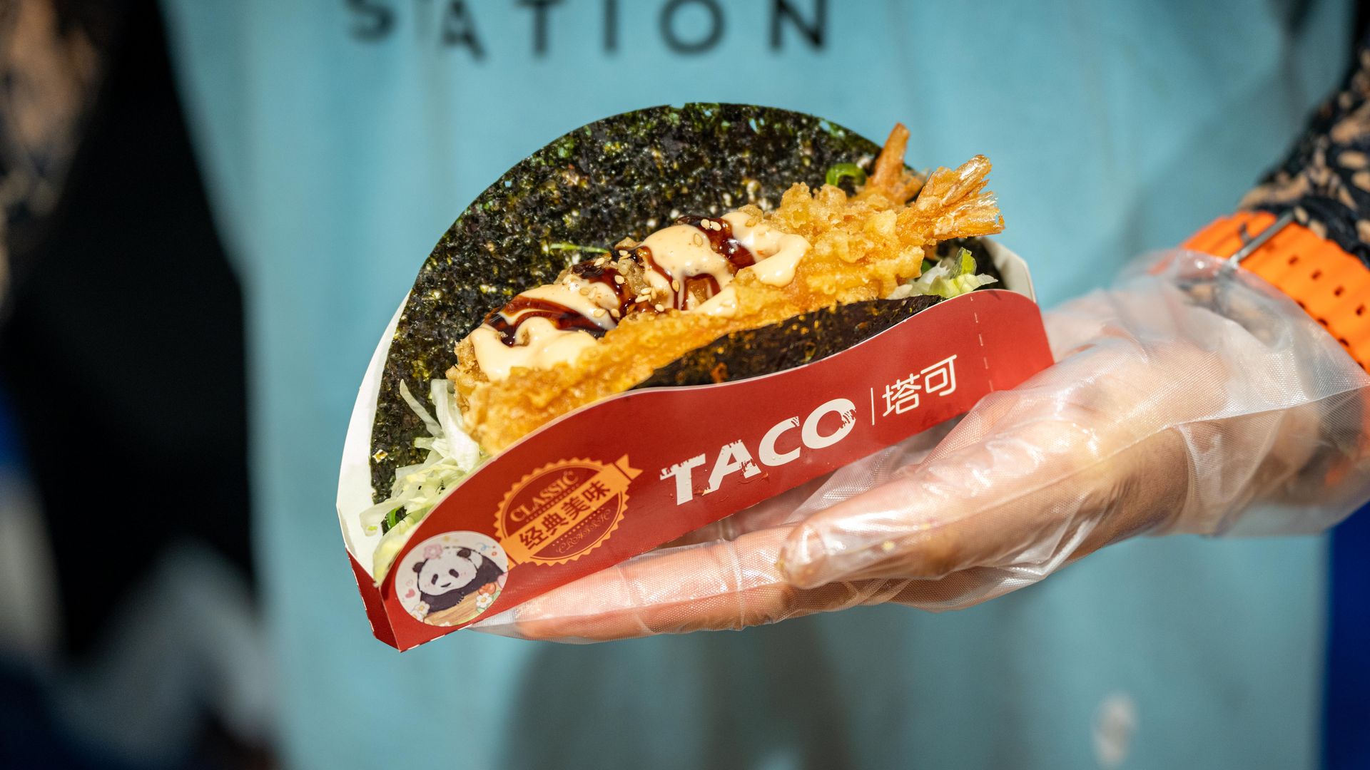Close-up of a fried shrimp taco with a seaweed shell, drizzled sauce, and shredded cabbage, held in a gloved hand. Red "TACO" wrapper with a panda sticker; blue shirt background.