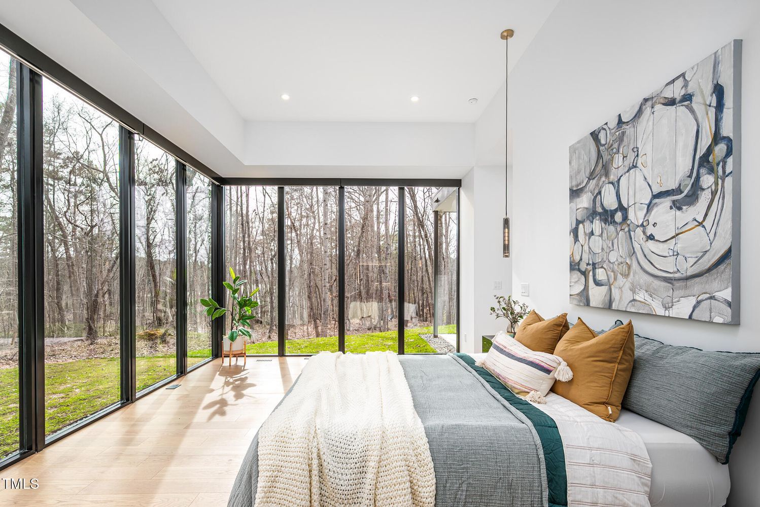 Modern bedroom with large floor-to-ceiling windows overlooking a wooded yard, light wood floors, gray bedding, brown and striped pillows, white knit blanket, abstract wall art, and a green plant.