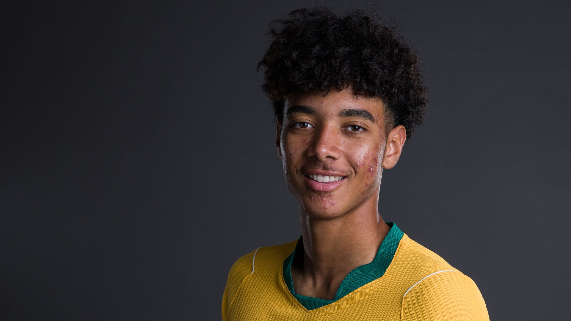 Young man with curly hair and acne, wearing a yellow sports jersey with green trim, smiles at camera against a dark gray studio background.