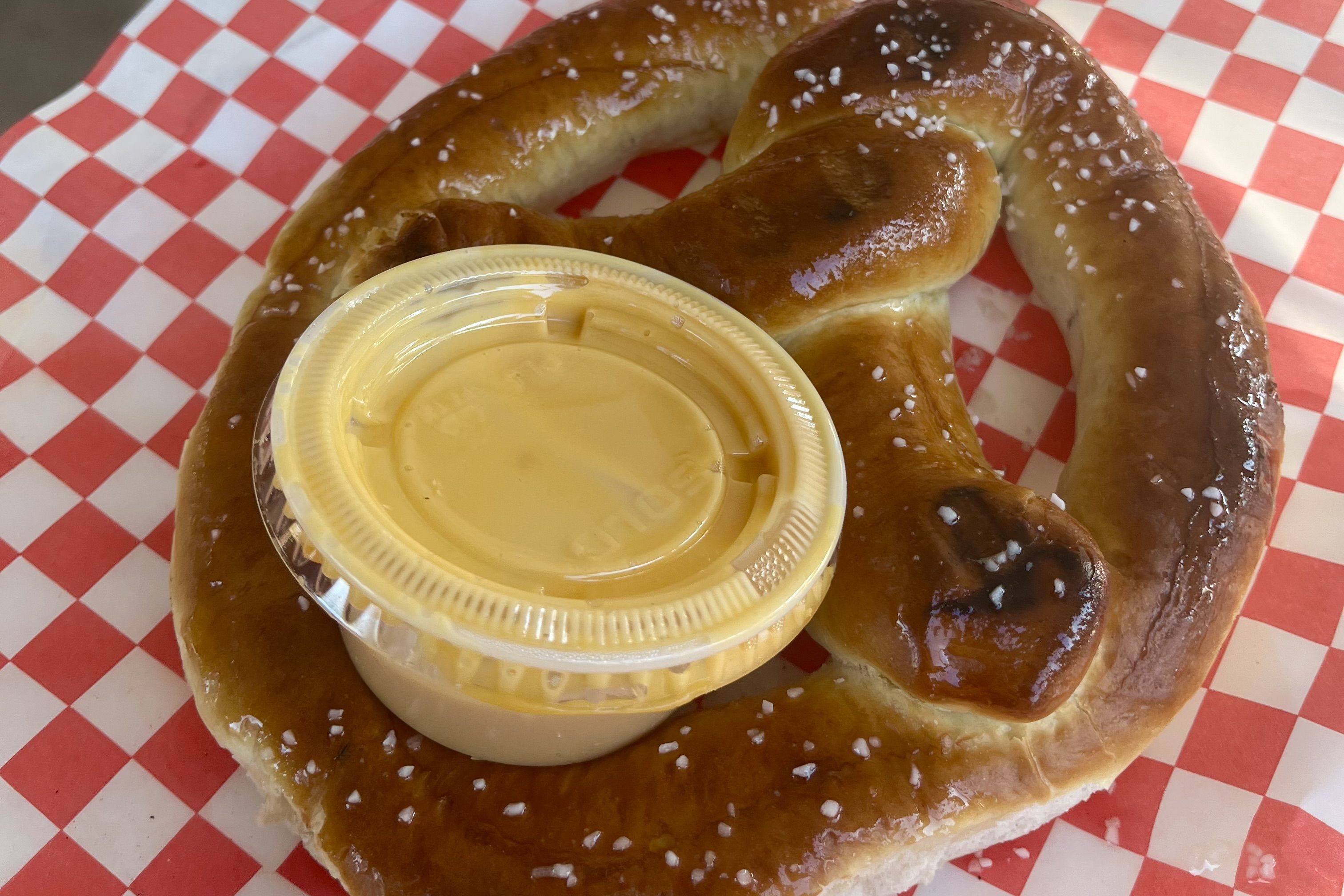 A glossy, salted soft pretzel twists on red-and-white checkered paper, with a yellow cheese dip in a plastic cup resting on its loop.