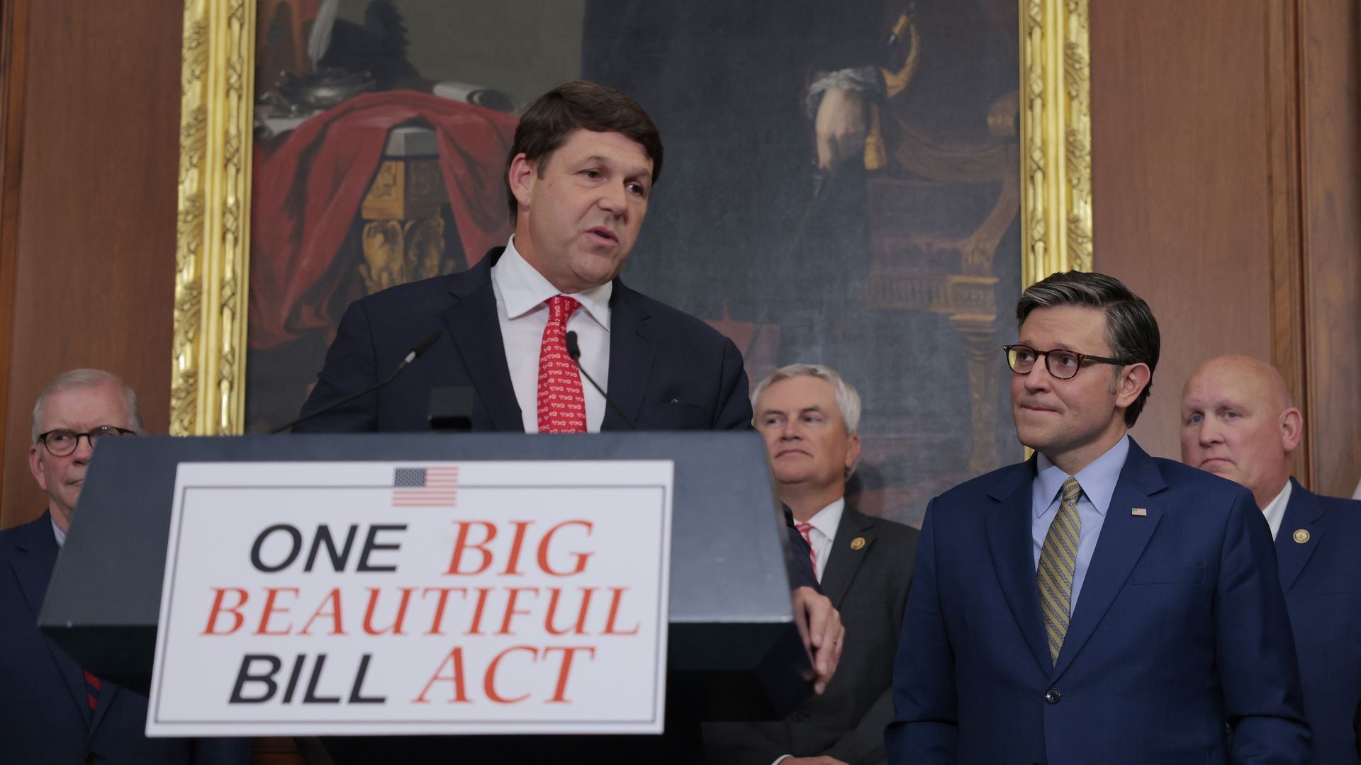 Man in a dark suit and red tie speaks at a podium, flanked by officials; a sign reads "ONE BIG BEAUTIFUL BILL ACT" in front of a large ornate painting.