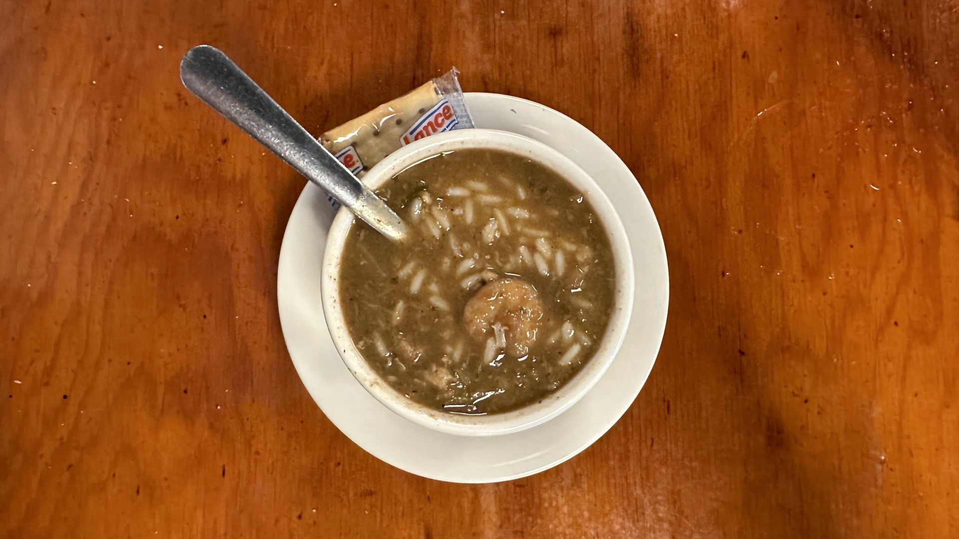 A cup of gumbo sits on a wooden table. Rice and shrimp are visible in the bowl, which has a silver spoon sticking out of it and the bowl rests on a plate with a cracket packet.