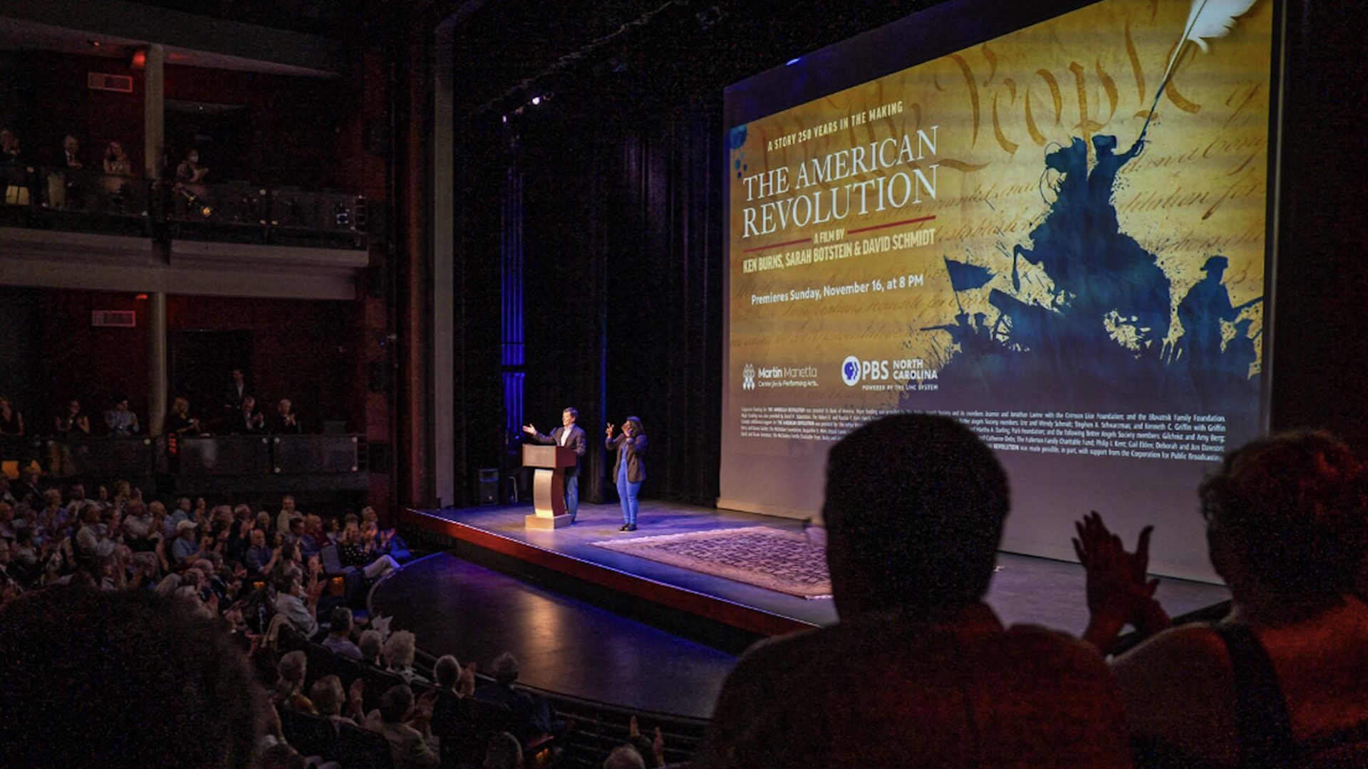 Audience applauding in a theater as a man at podium and a woman sign language interpreter stand on stage beside a large screen showing "The American Revolution" PBS film premiere details.
