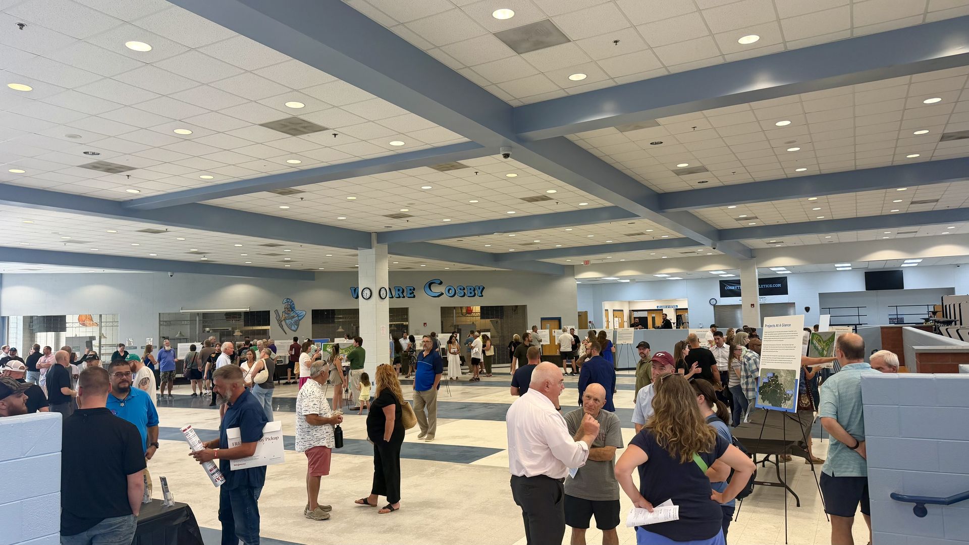 Crowded indoor exhibition hall with a white grid ceiling and blue support beams. People mingle among booths and displays, holding papers and chatting, with light blue walls in the background.