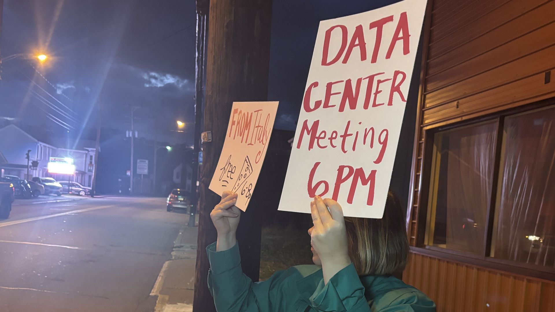 Person in teal jacket holding signs reading "DATA CENTER Meeting 6 PM" and "FROM Italy free" with pizza slice illustration, standing on dark street with lit buildings and cars at night.