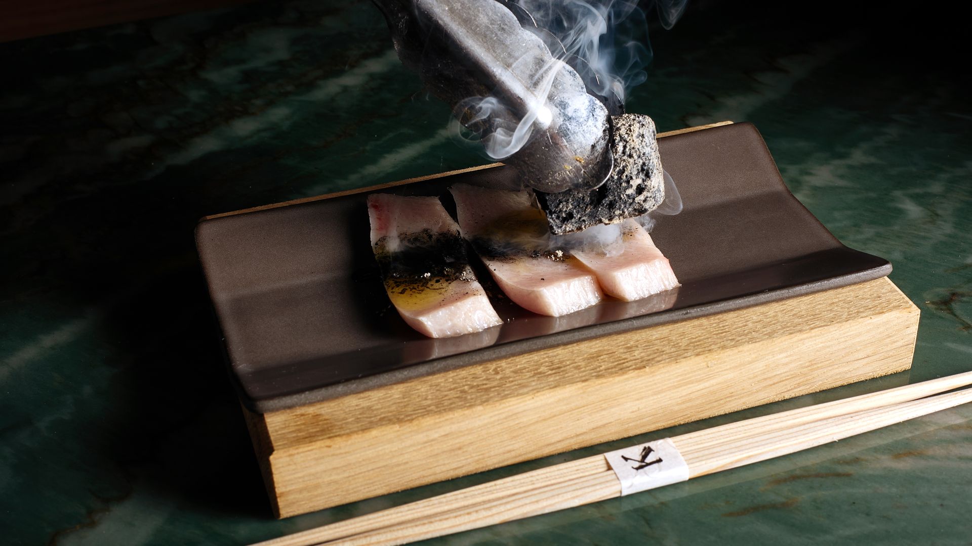 Close-up of a metal charcoal torch pressing a glowing ember onto salmon slices on a wooden board; smoke swirls upward against a dark background.