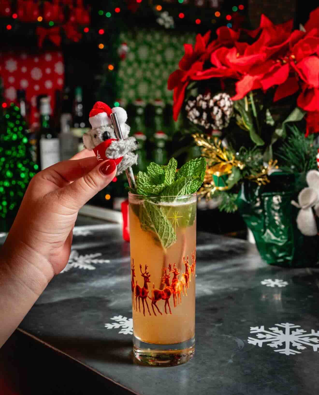 Hand holding a metal straw with a small koala wearing a red Santa hat, above a tall festive drink garnished with fresh green mint in a glass decorated with a red reindeer pattern, on a table with snowflake designs.