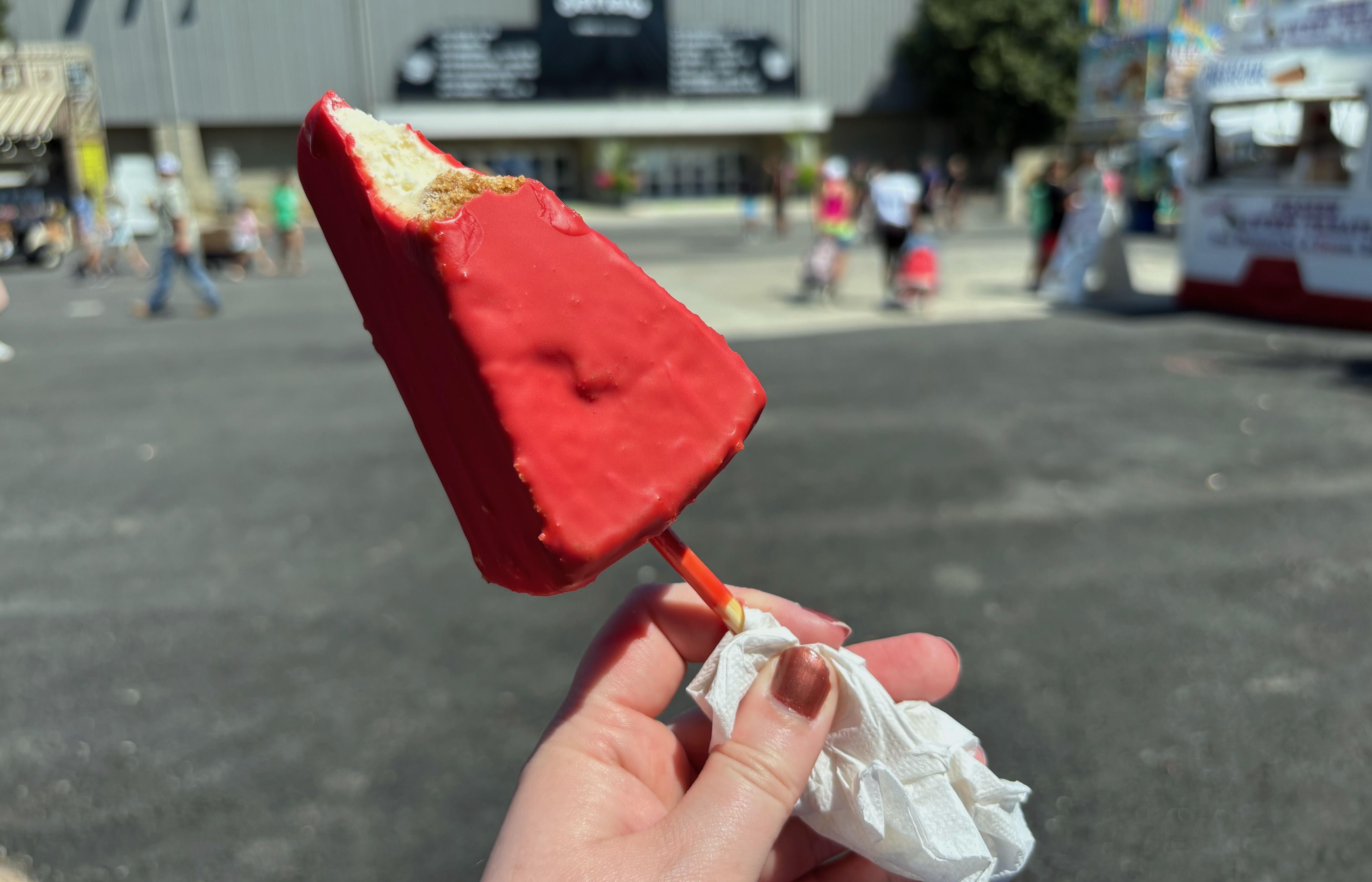 Hand holding a partially eaten red-coated frozen cheesesteak on a stick, with a white napkin wrapped around the stick, in a sunny outdoor setting with blurred people and buildings in the background.