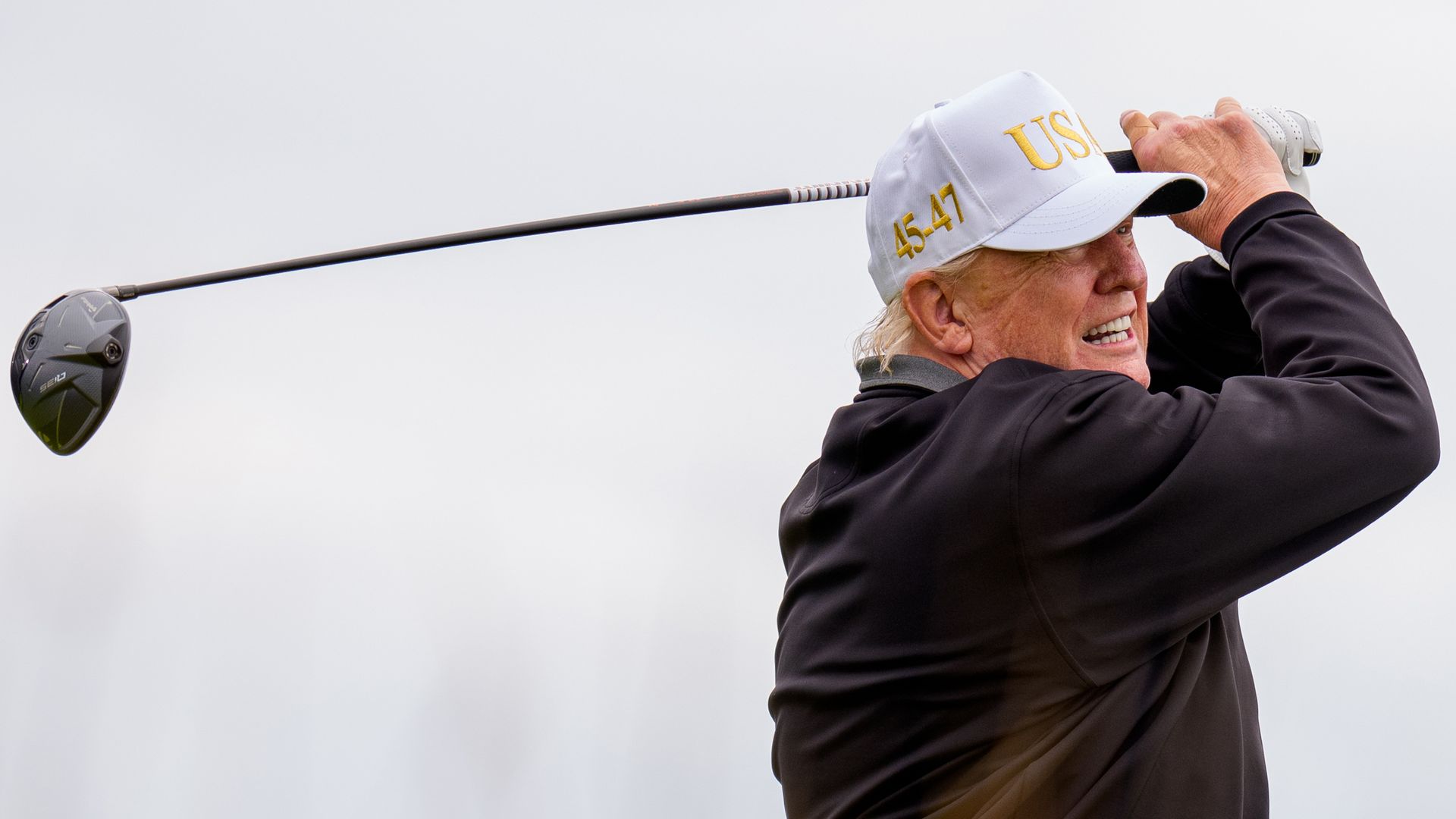 A person in a black long-sleeve shirt and white hate swings a golf club
