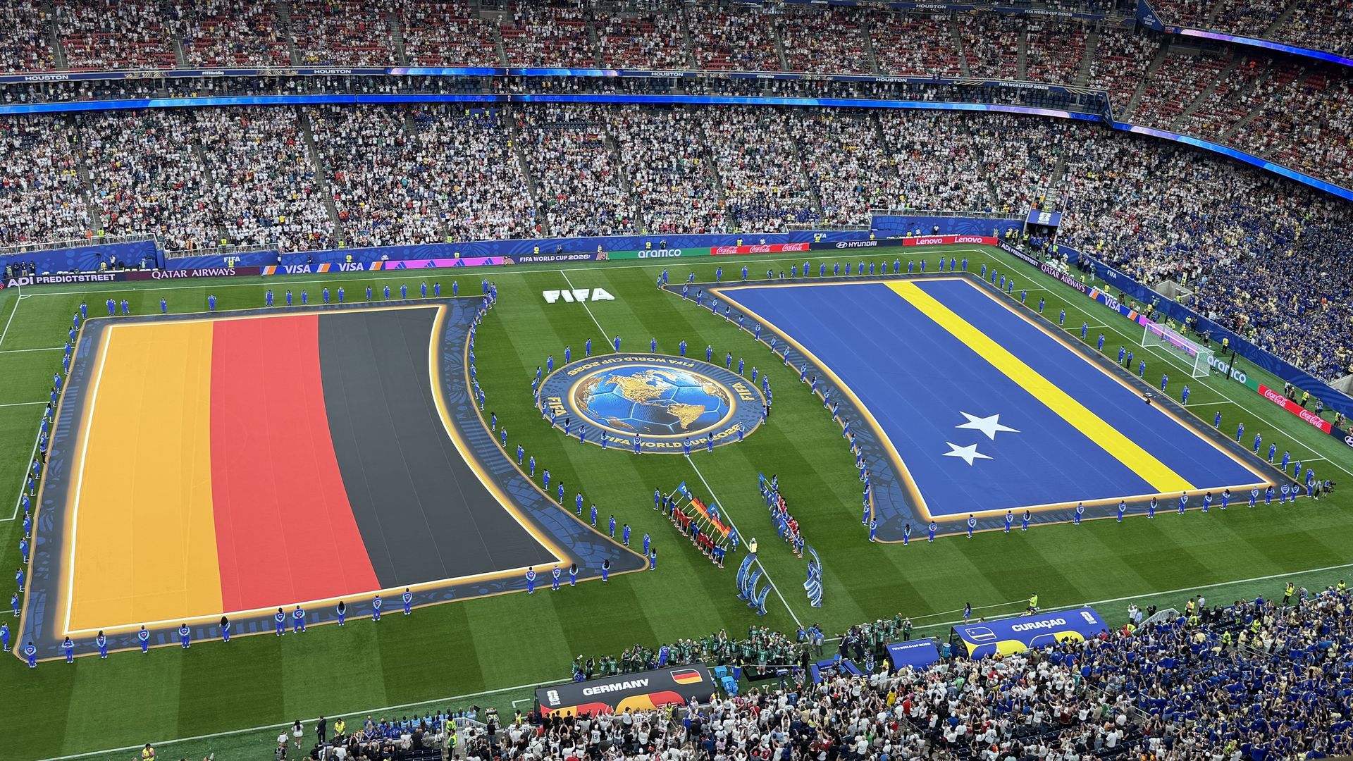 Panoramic view of a packed stadium for FIFA World Cup 2026 in Houston. Crowds fill the stands as blue-clad players line up on a colorfully bordered field with a central globe emblem.