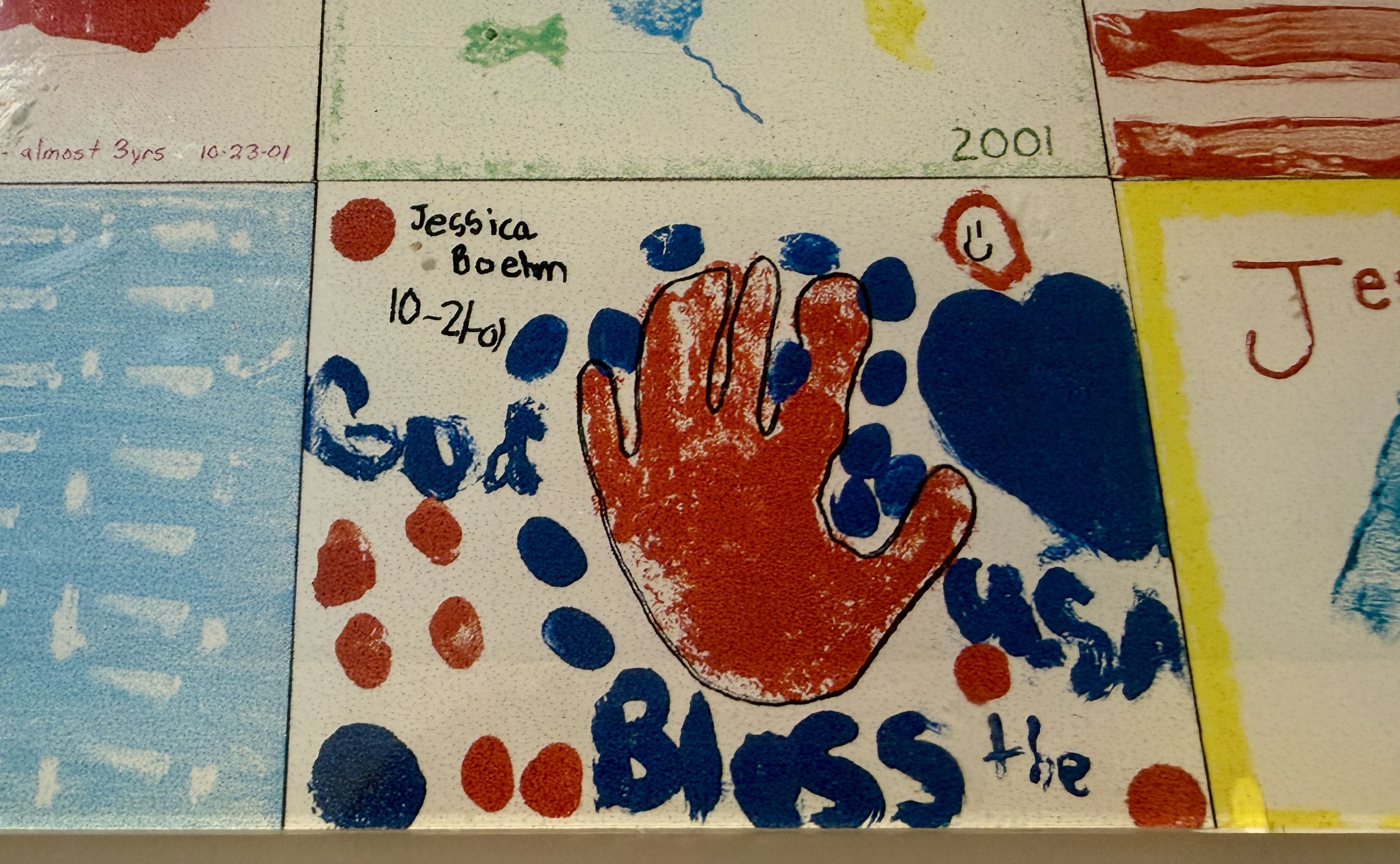 Close-up of a mosaic tile featuring a red handprint with blue dots around it, handwritten text "Jessica Boehm" and a date; a green "2001" tile sits above amid blue, red, and yellow blocks.