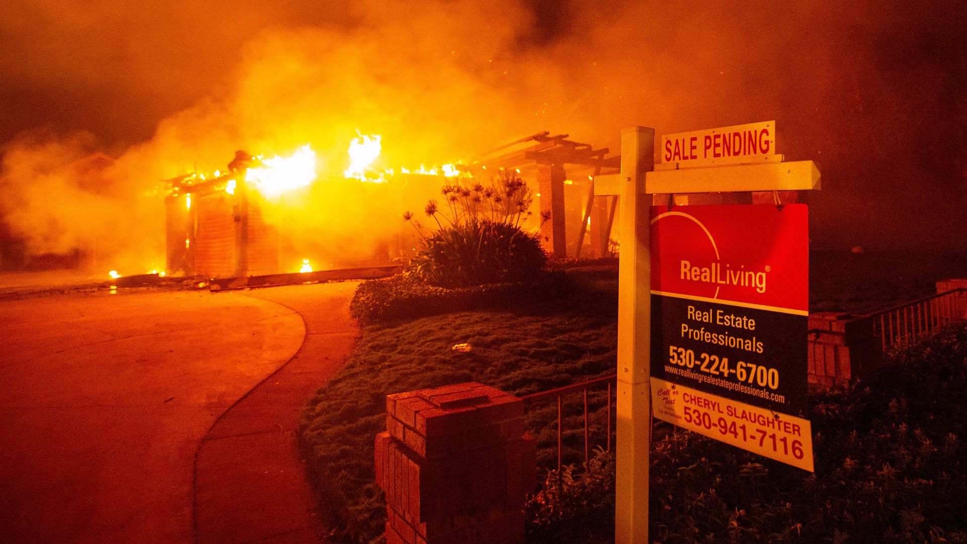 A real estate sign sits in front of a burning structure in Redding, California. 
