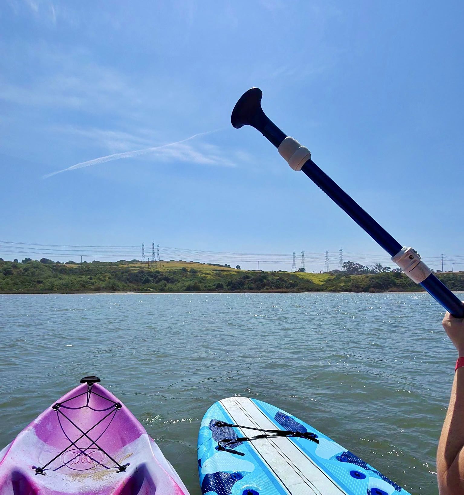 Two colorful stand-up paddleboards float on calm water; a person raises a black, curved-blade paddle. In the distance are green hills with power lines, under a bright blue sky.