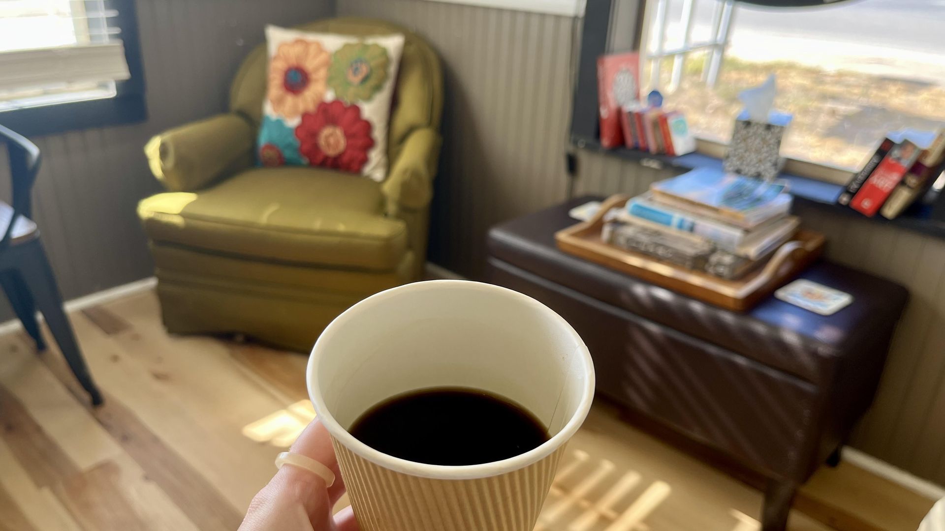 A black cup of coffee in front of a light-filled window with books and a plush green chair.