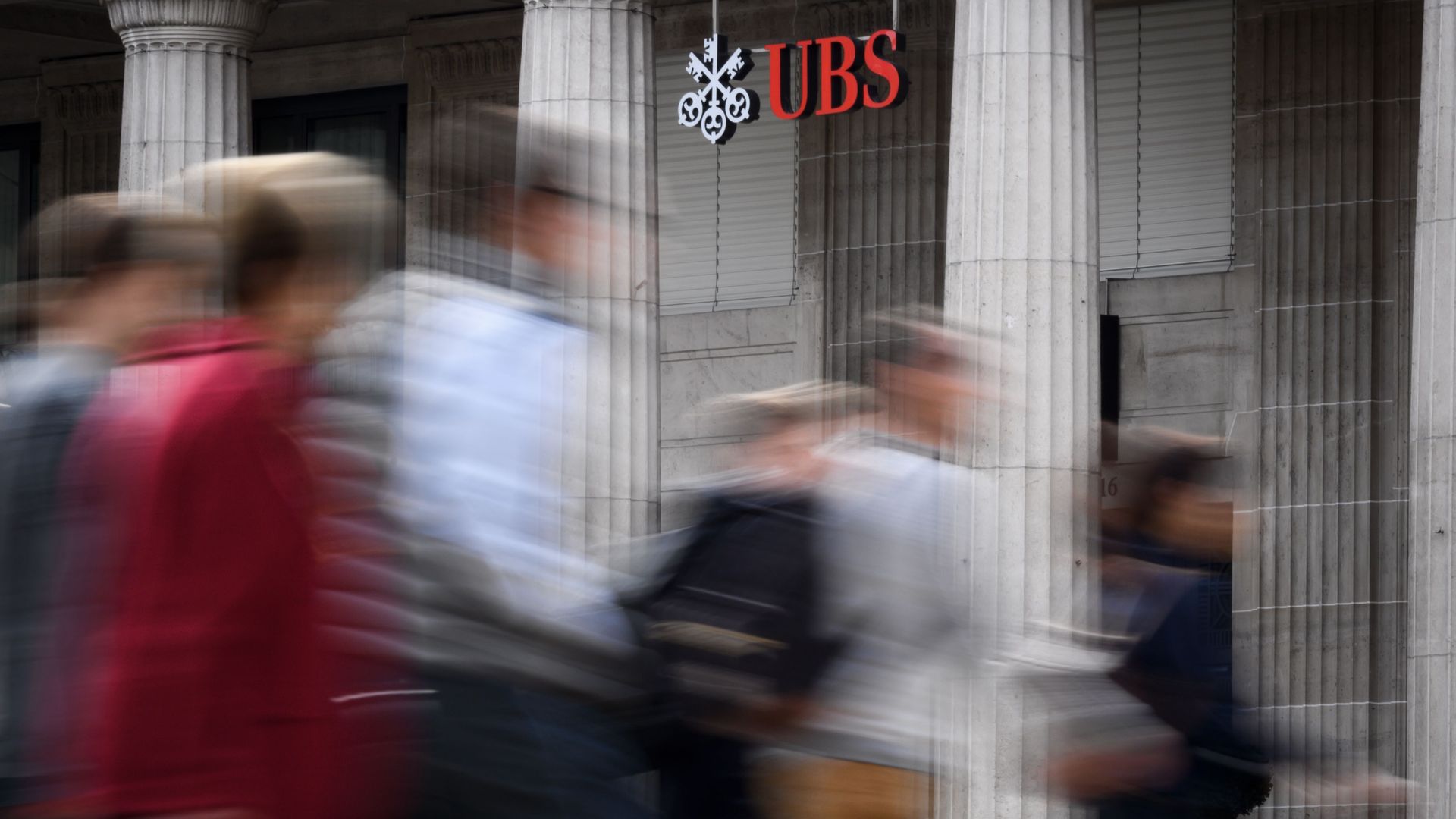 People cross a road past a sign of Swiss banking giant UBS hanging outside a branch in Lausanne on October 6, 2018.