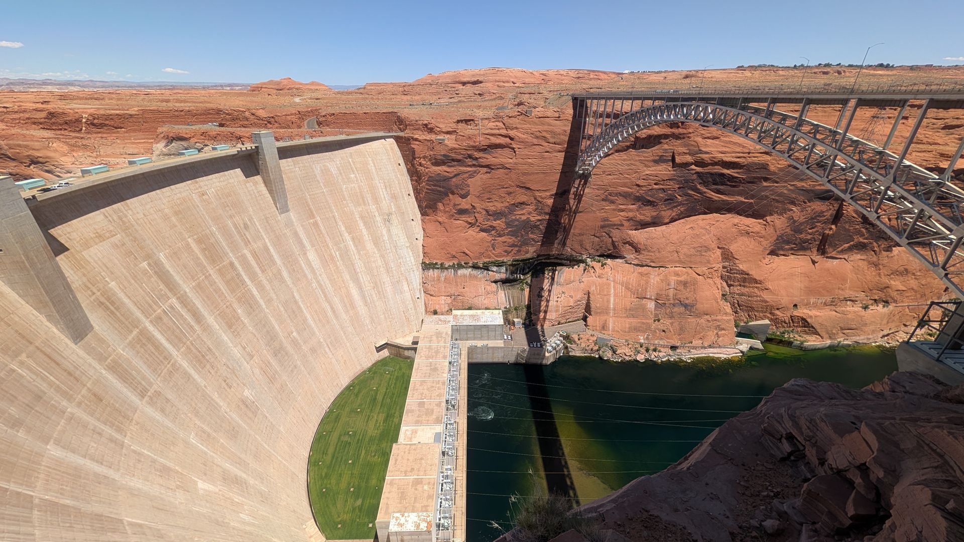 Massive concrete dam curves along a red sandstone canyon with a green reservoir at its base; a steel arch bridge spans the gorge to the right under a clear blue sky.