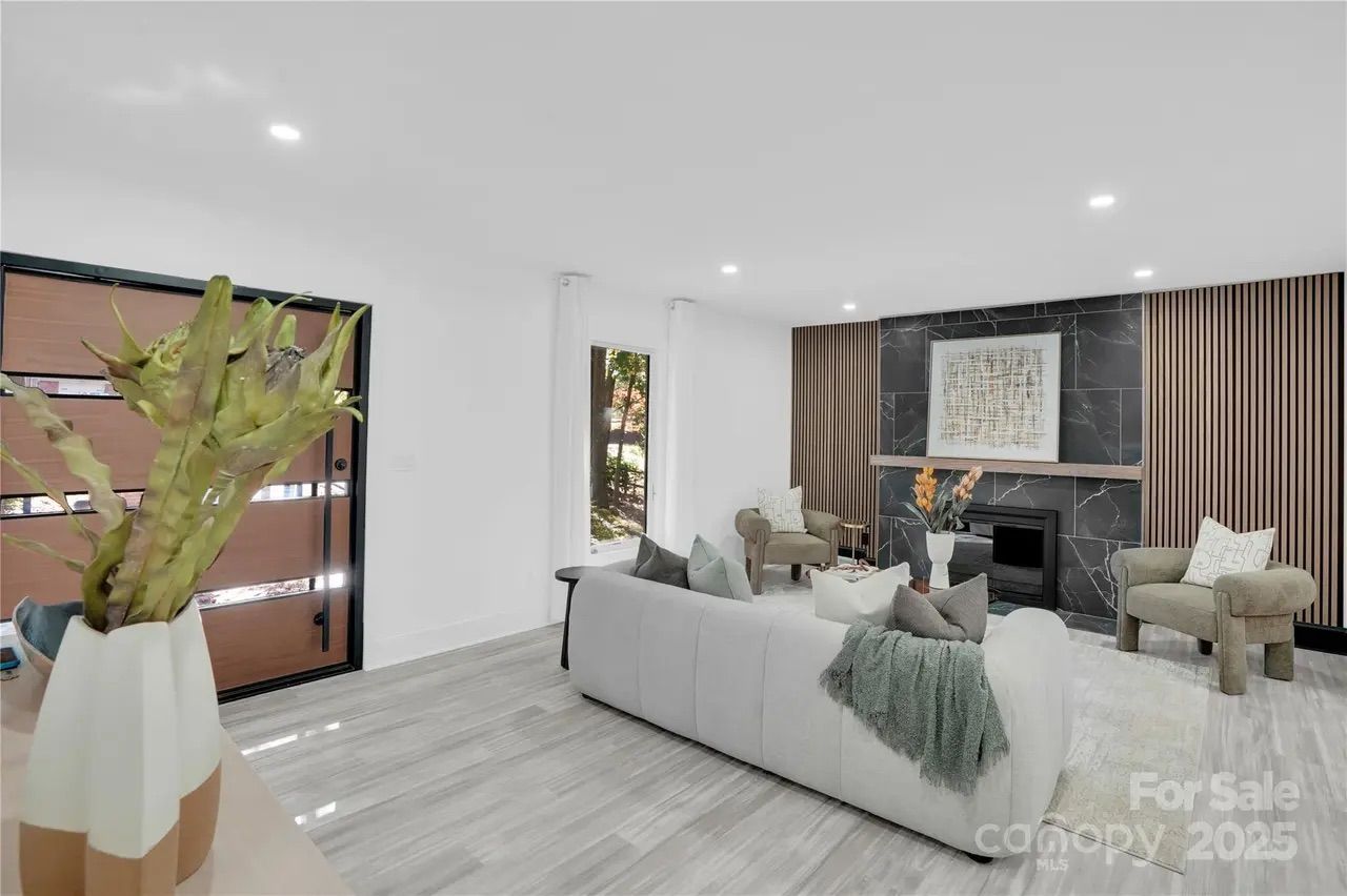 Modern living room with white walls, light gray wood floor, beige couch with cushions and throw, brown armchairs, black marble fireplace with abstract art, and wooden accent panels.