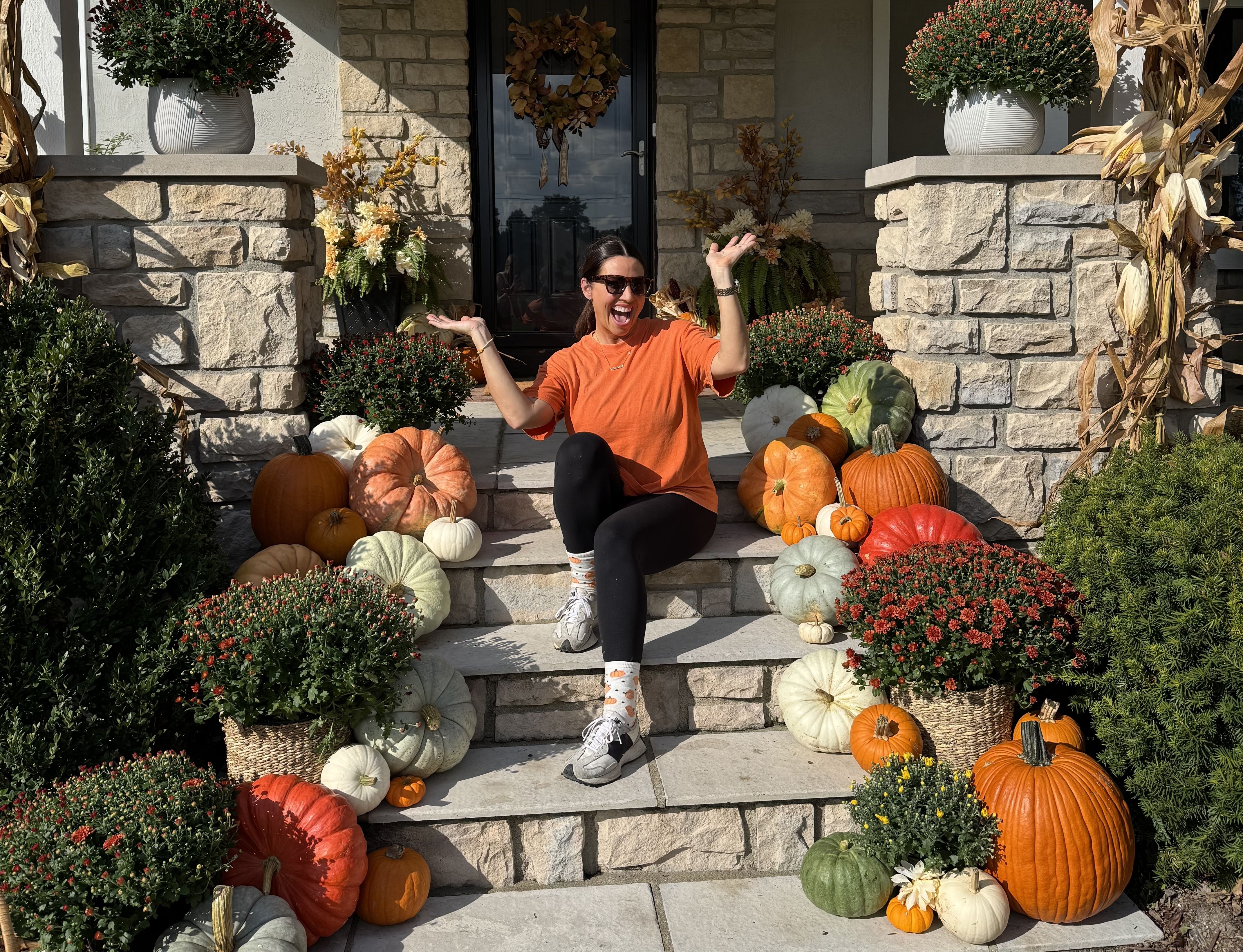 Woman in orange shirt and black pants sitting on stone steps surrounded by various pumpkins and mums in pots, with autumn decorations on a house porch.