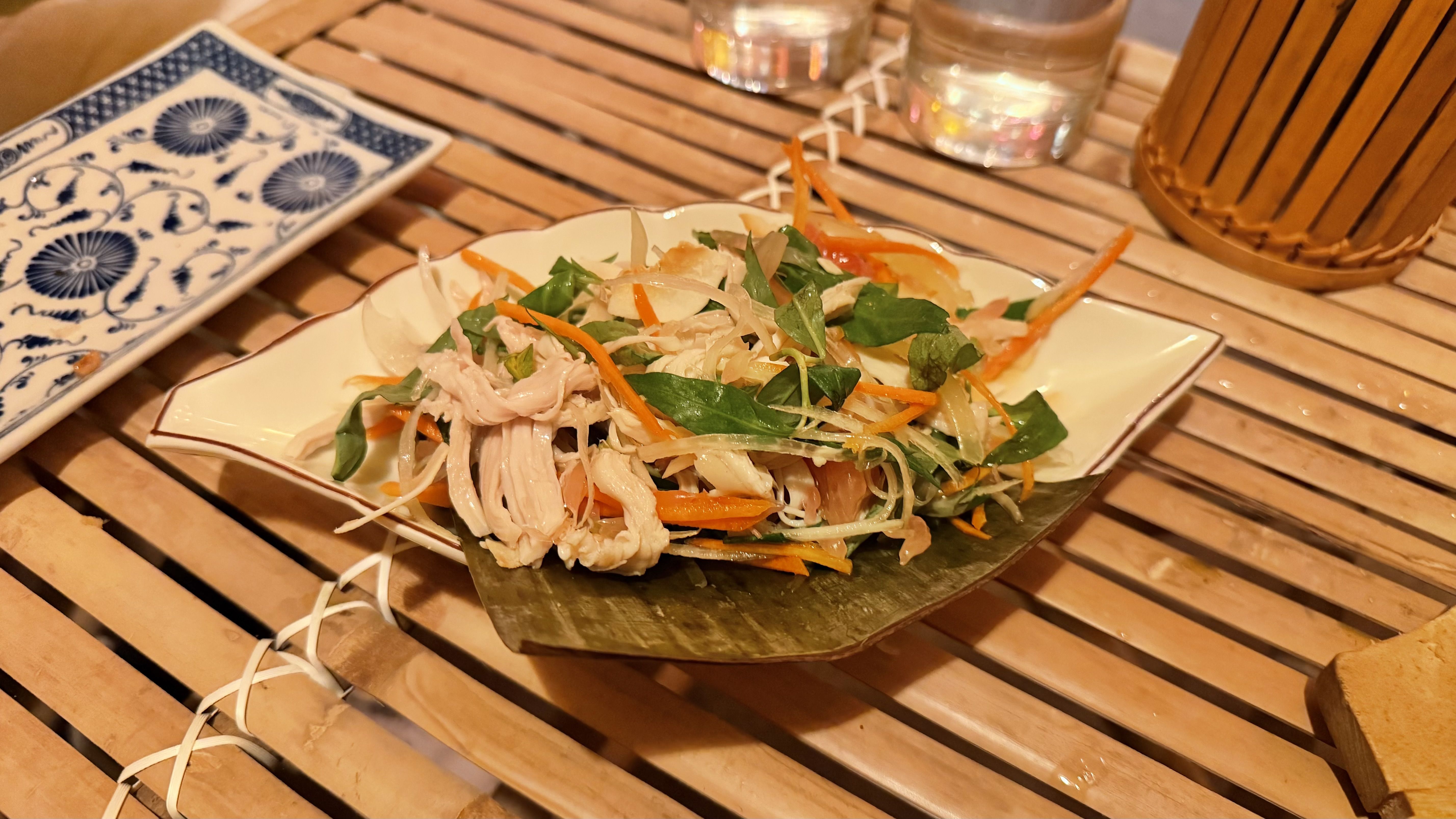 Plate of shredded chicken salad with green leafy herbs, thin carrot and onion strips on a leaf-shaped dish on a wooden table, next to a blue and white patterned plate.