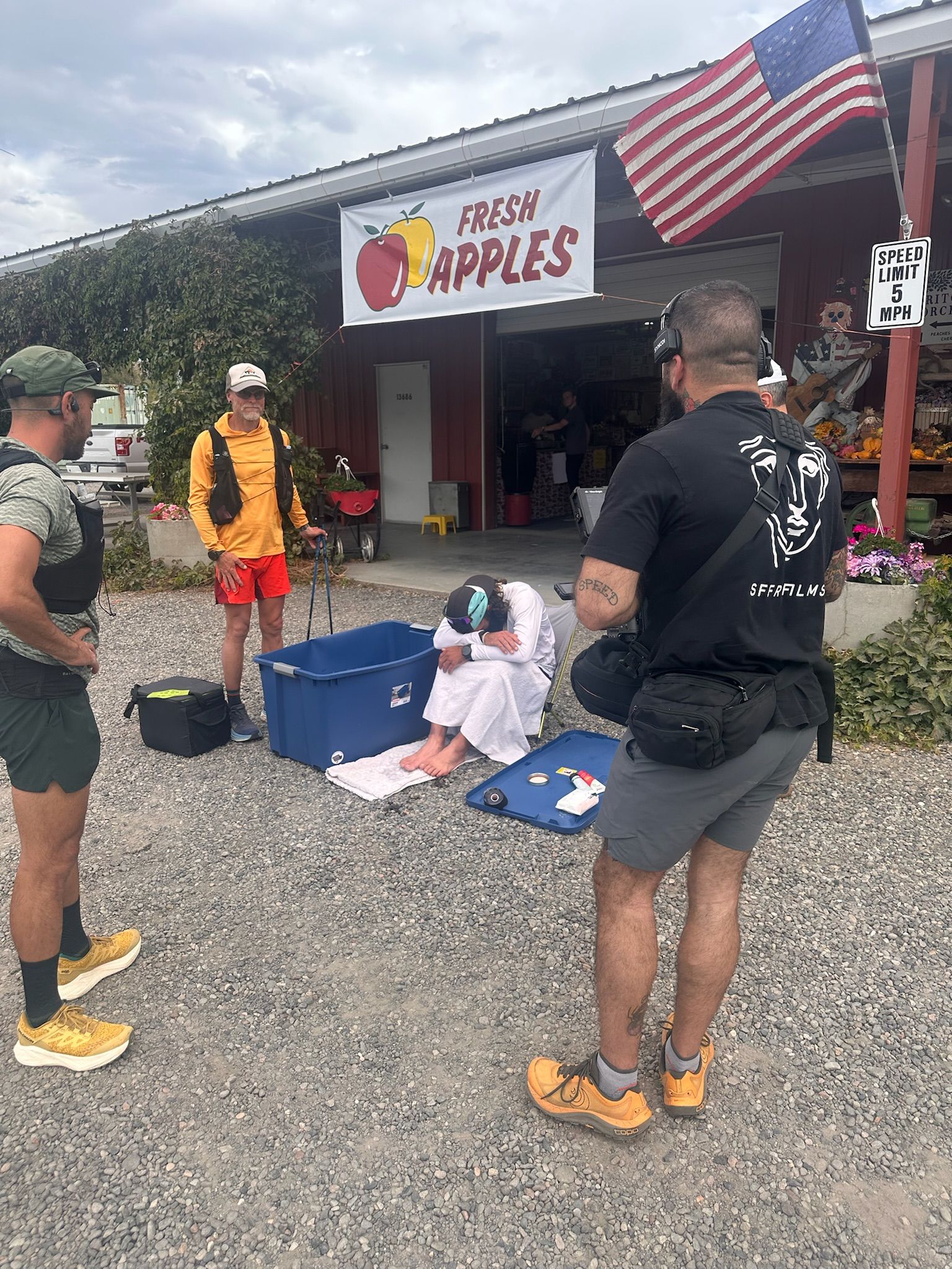 Outdoor market in front of a red storefront with a white banner reading Fresh Apples and apple graphics. An American flag hangs overhead as several people in athletic gear stand near a blue bin.