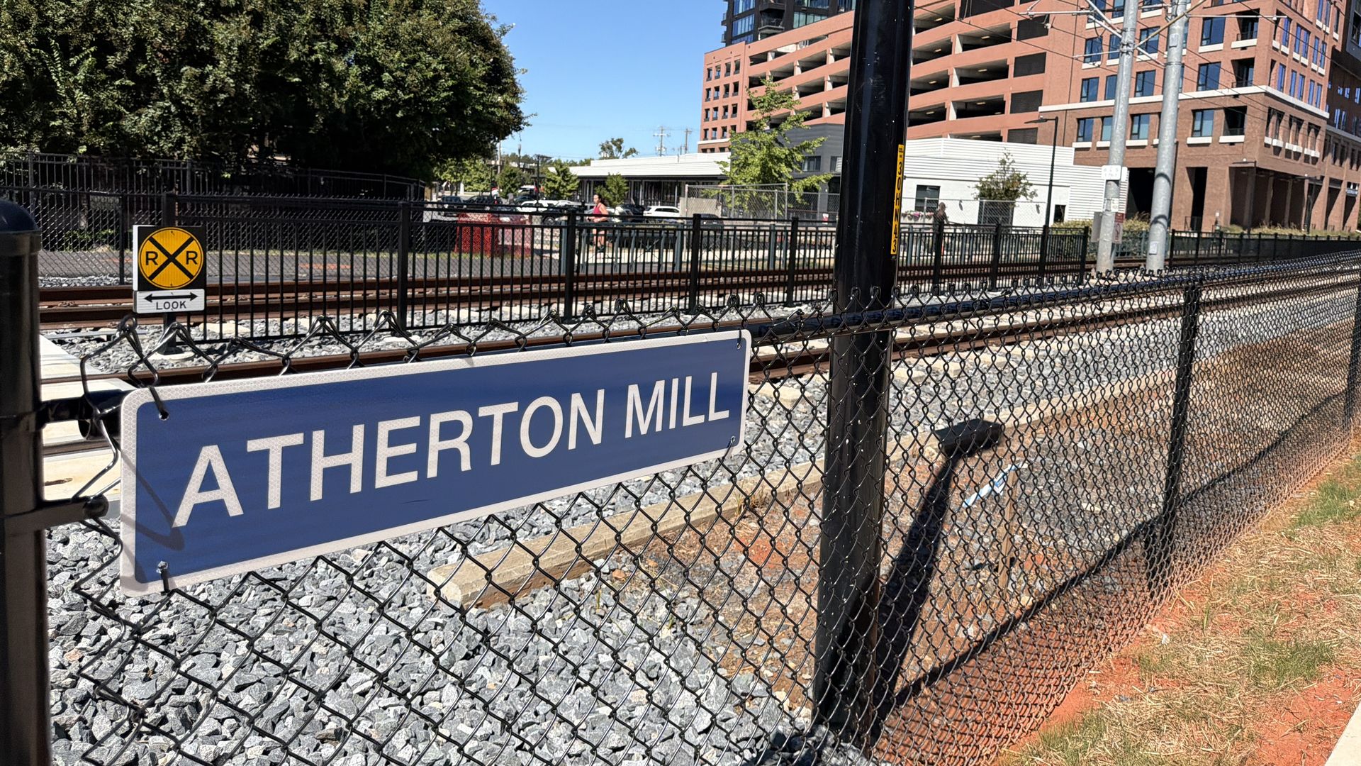 Train tracks at Atherton Mill station with a blue Atherton Mill sign on a chain-link fence, a tall brown brick building, overhead wires, and a few people under a clear blue sky.