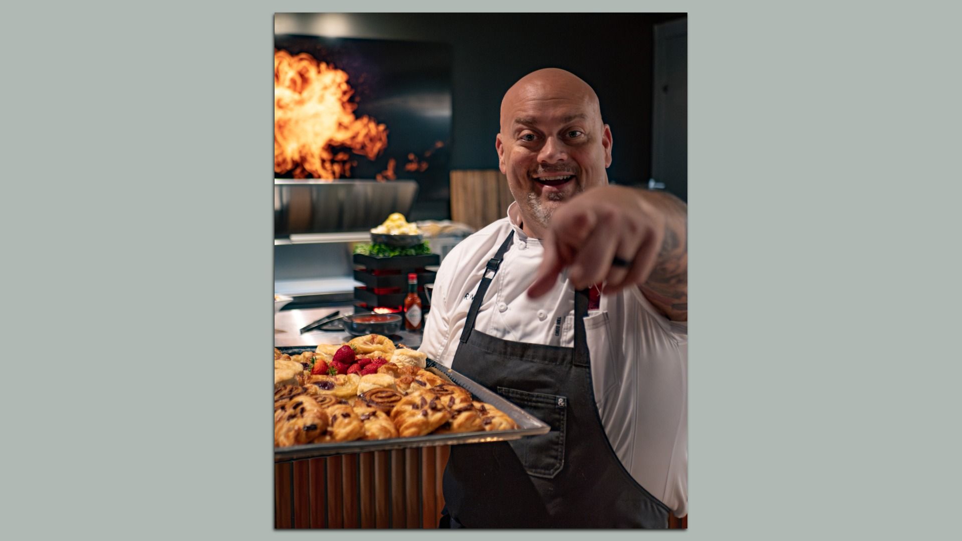 Cheerful bald chef in a white shirt and black apron presents a tray of pastries toward the camera in a busy kitchen, flames blazing in the background.