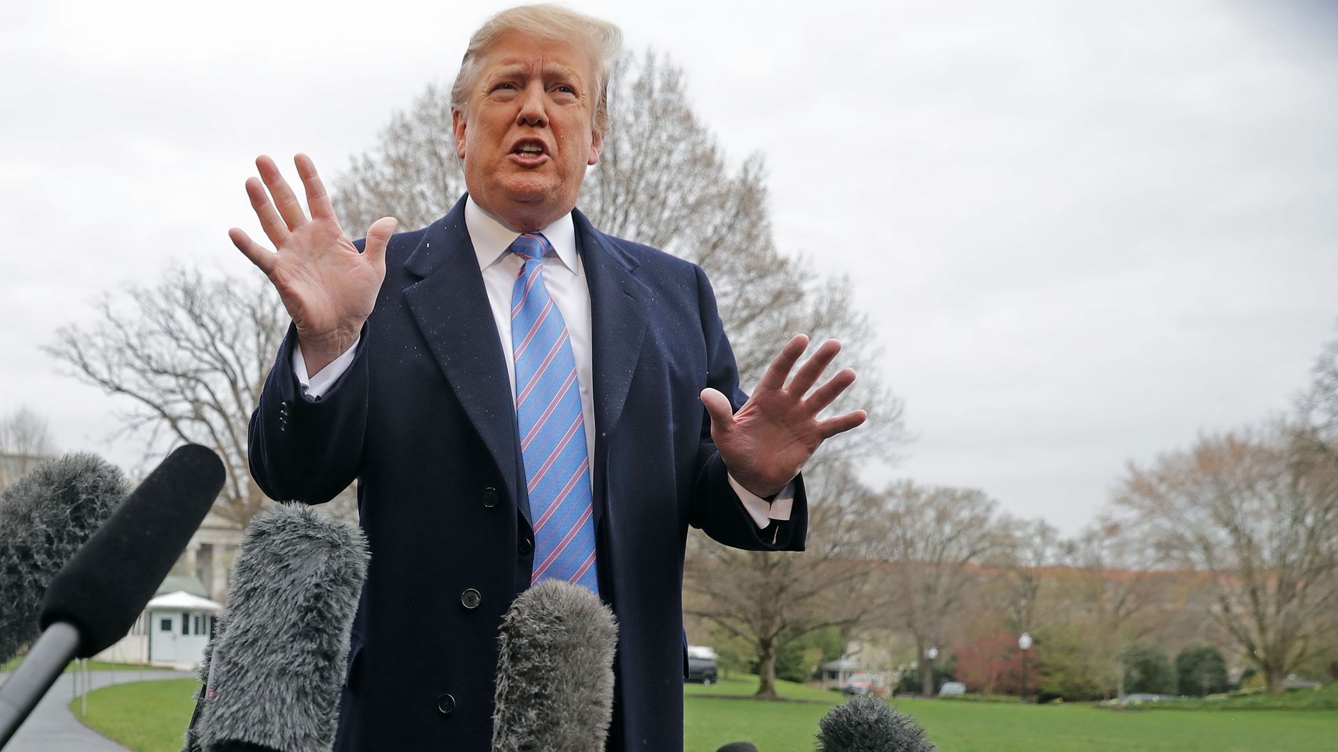 In this image, Trump speaks with both hands open in front of boom mics and reporters on the White House lawn.