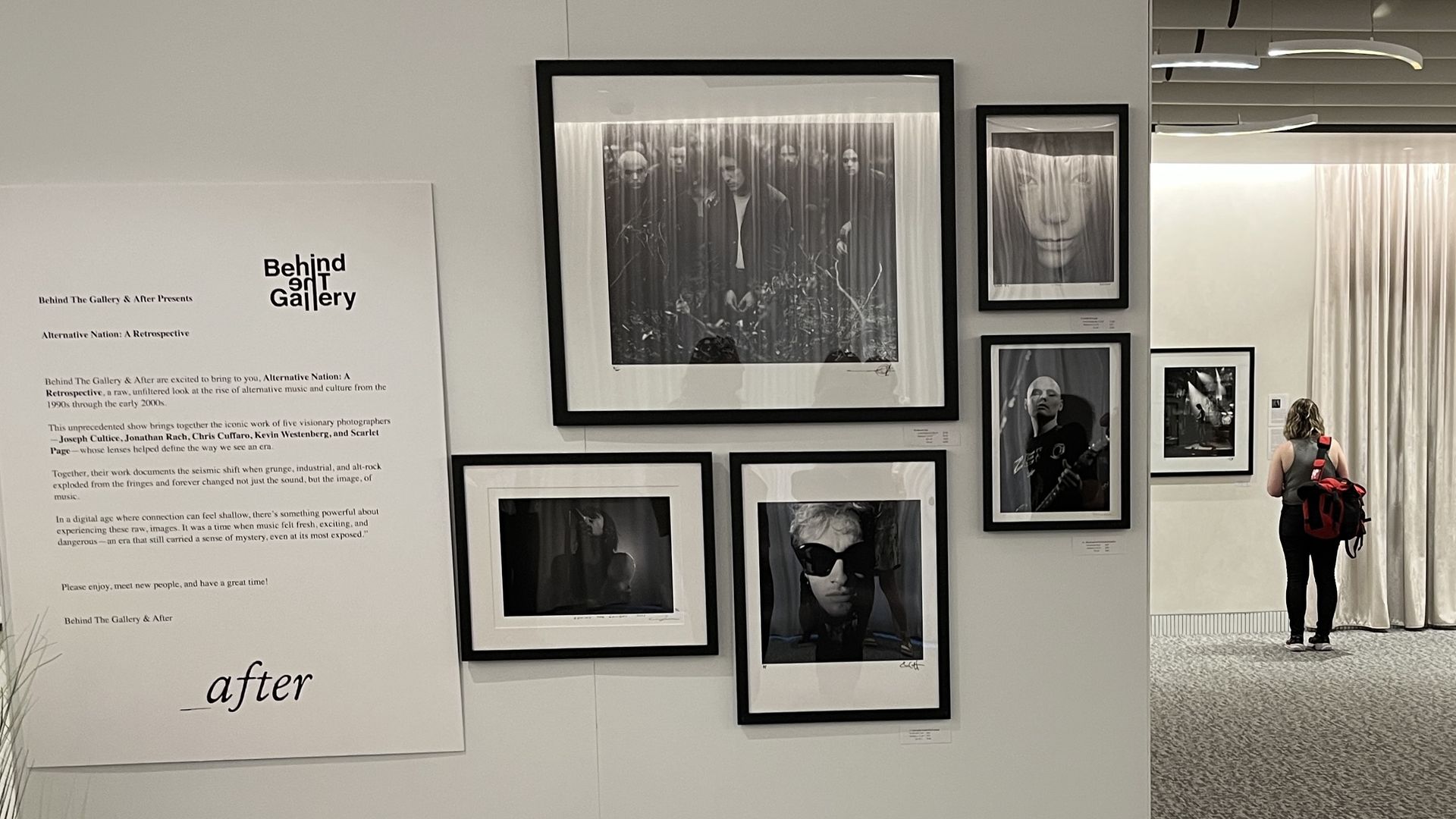 Wall exhibit titled "Behind The Gallery" showing black-and-white photos from "Alternative Nation: A Retrospective" featuring portraits and musicians, with a woman viewing art in gallery.