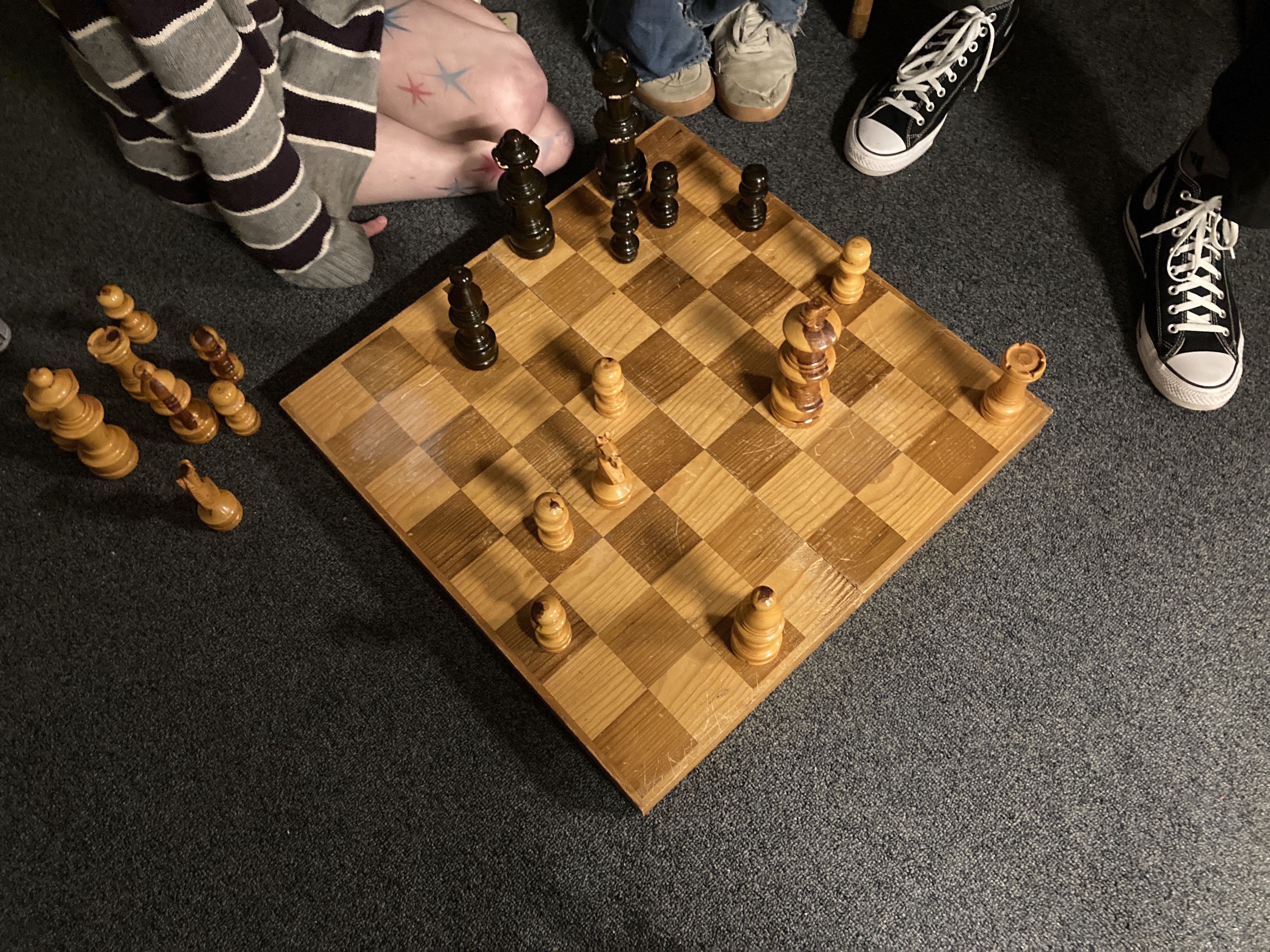 The knees and feet of three people around an oversized wooden chess board on a grey carpeted floor.