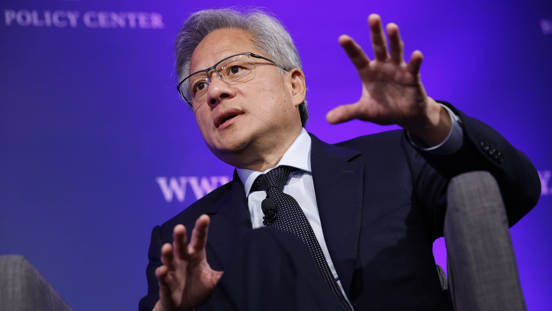 photo of a man with gray hair in a suit and glasses sitting on stage against a purple background and talking with his hands open